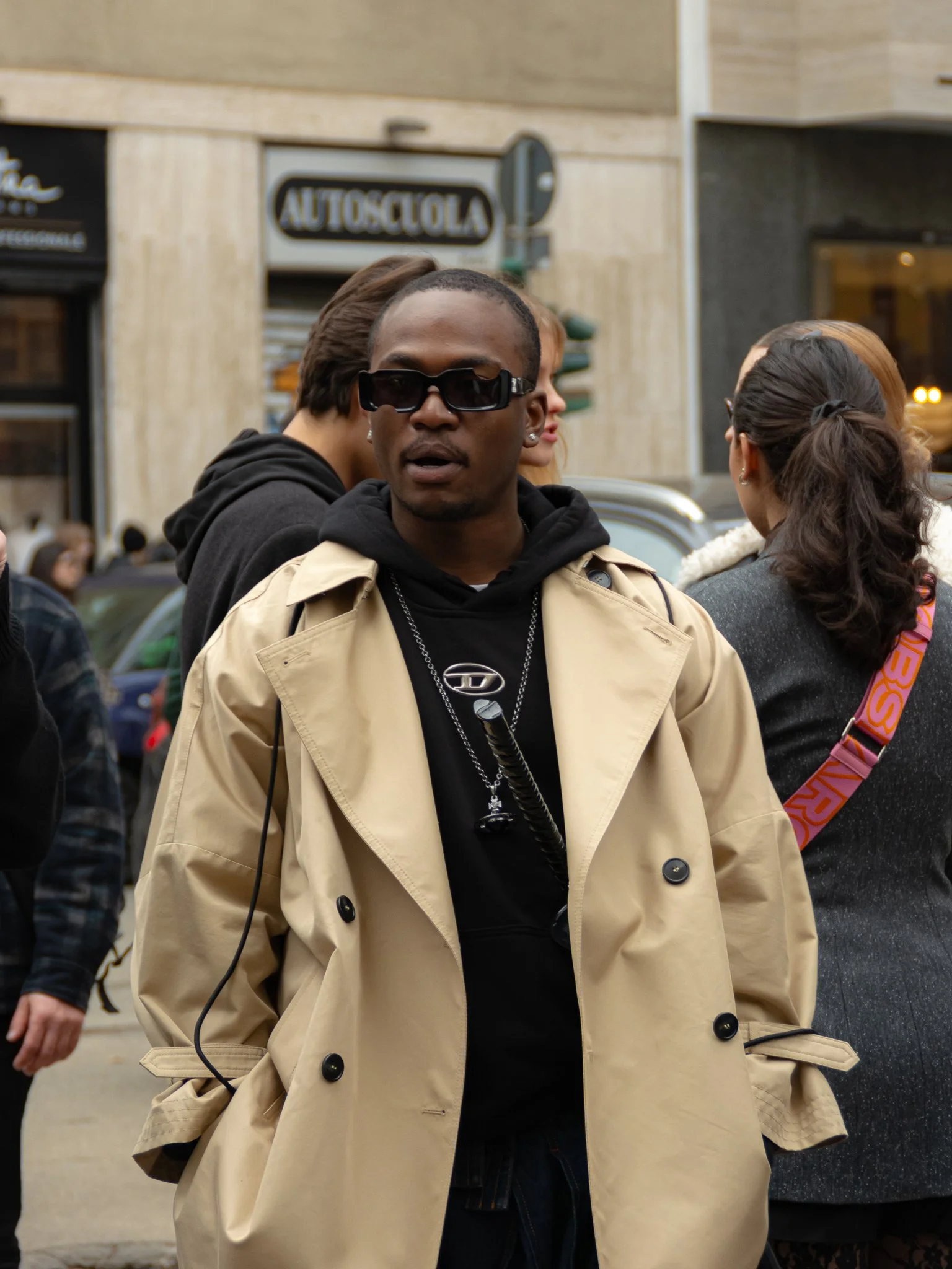 Man wearing a beige trench coat and black sunglasses standing in a busy street with people in the background.