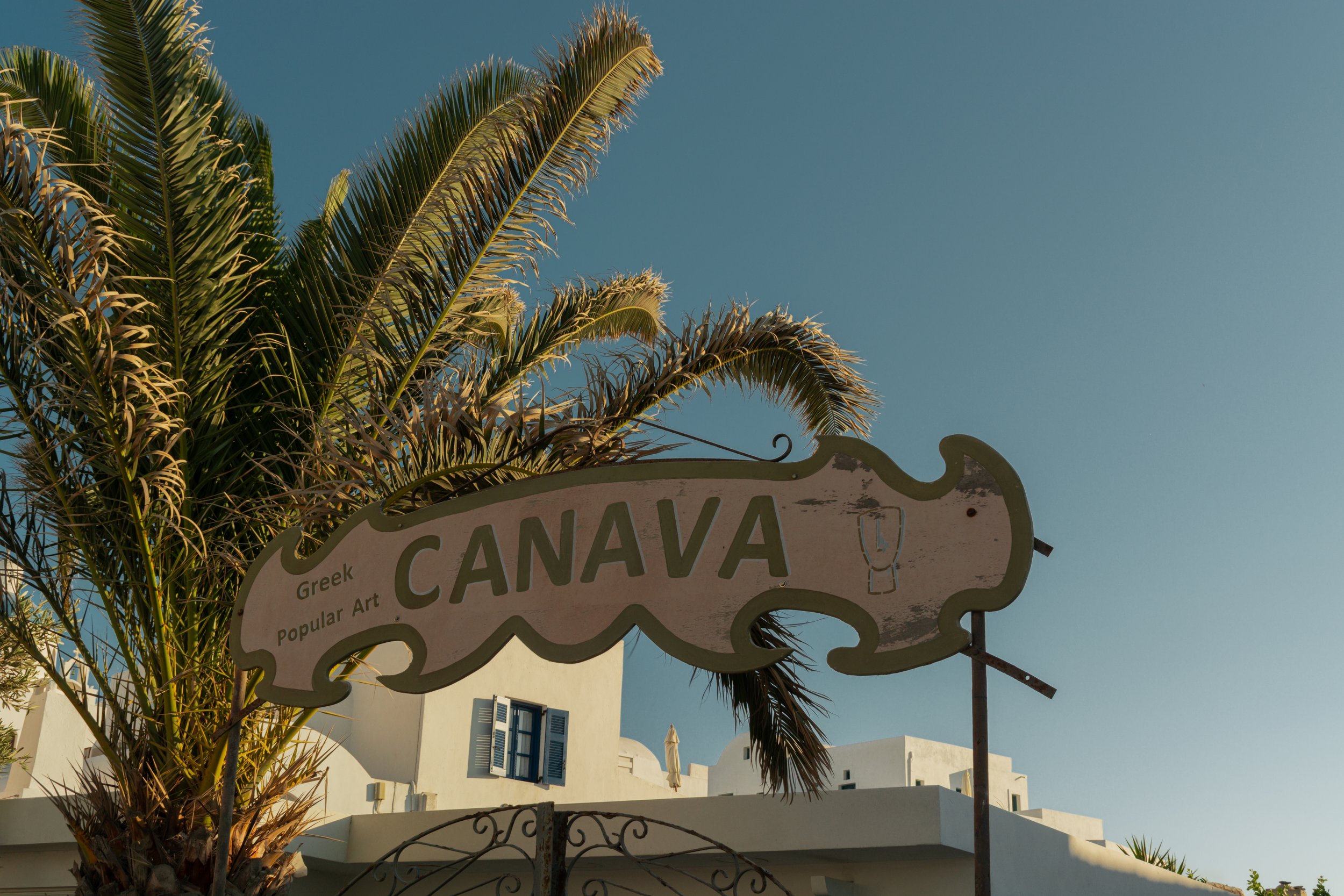 Vintage sign reading 'CANAVA Greek Popular Art' near palm tree and white building, clear blue sky.