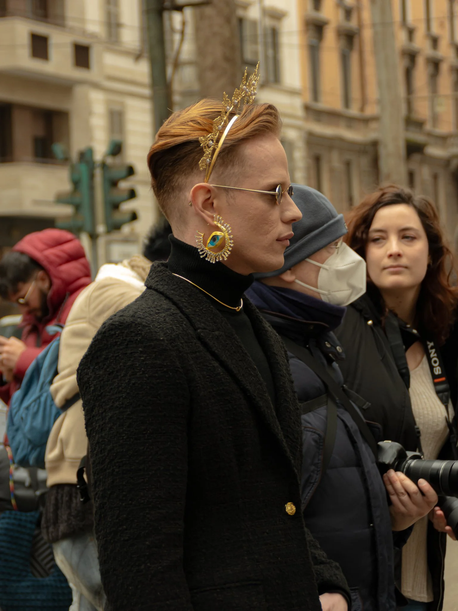 A person with short hair, wearing a gold crown and a black coat, standing among a crowd. They have an ornate earring and sunglasses. People around them are wearing winter clothing, and one person is holding a camera. Urban background with buildings a