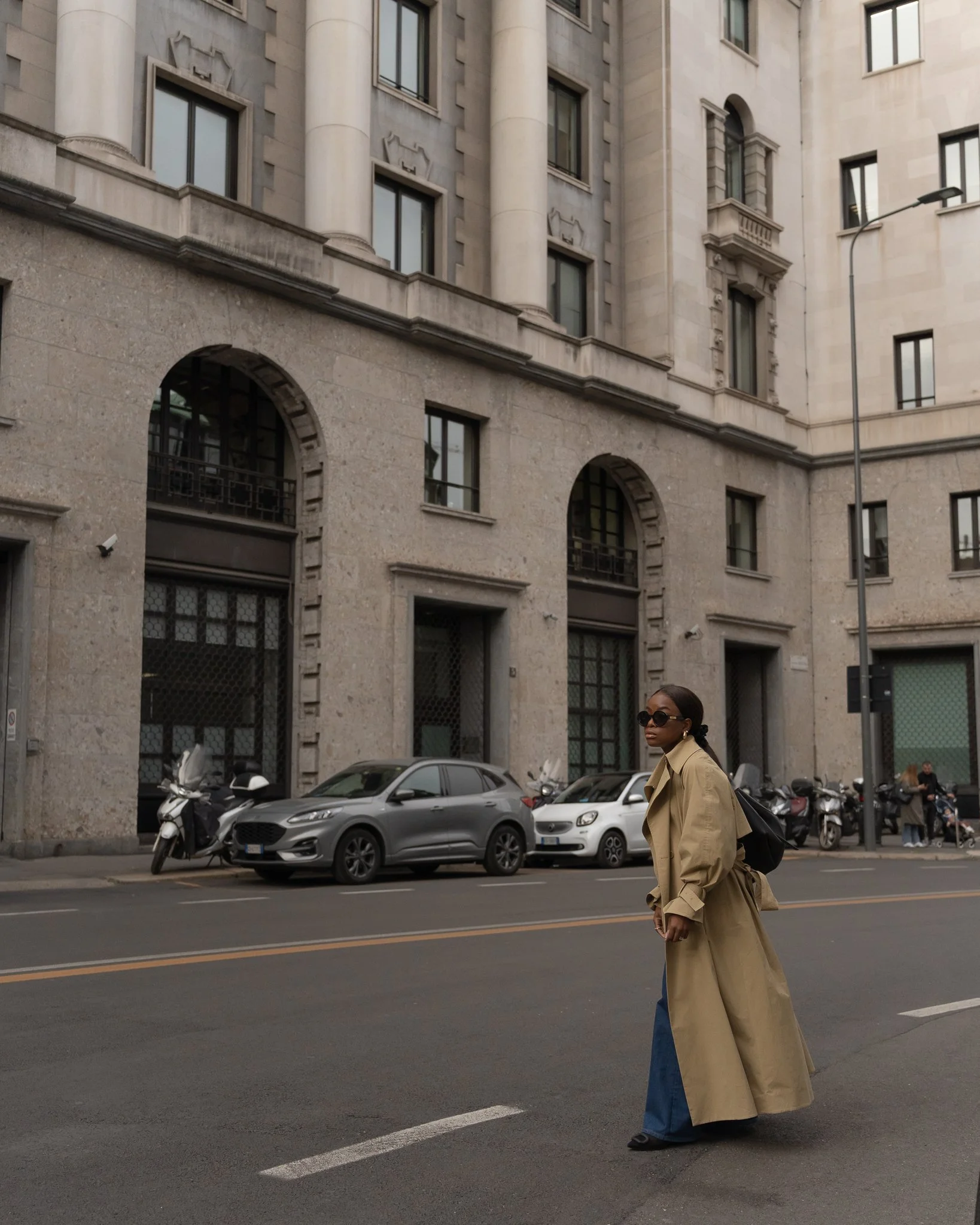 Person wearing a long beige trench coat and sunglasses walking across a city street, with a classic European building and parked vehicles in the background.