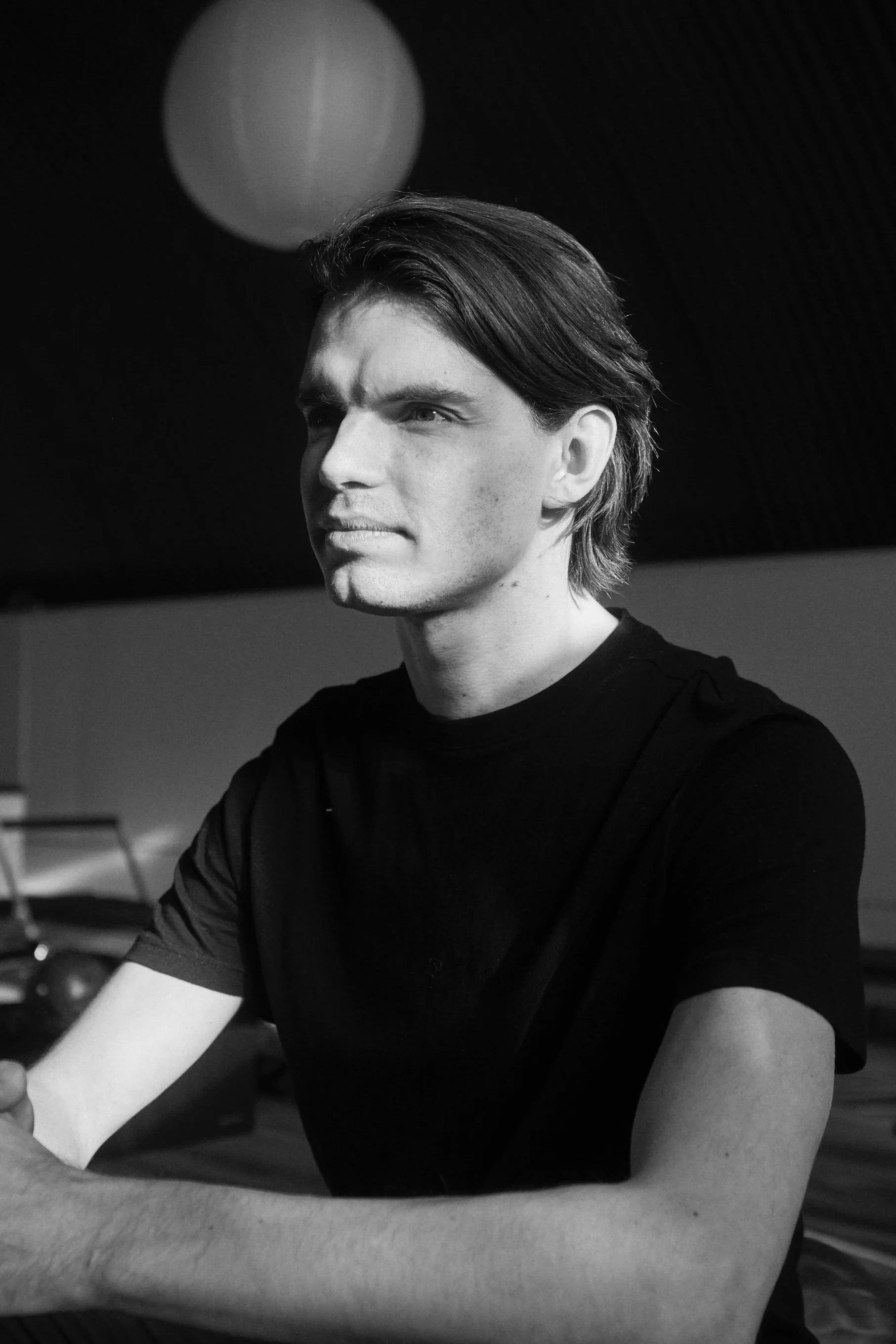 Black and white photo of a young man with medium-length hair, wearing a black t-shirt, looking slightly to the side.