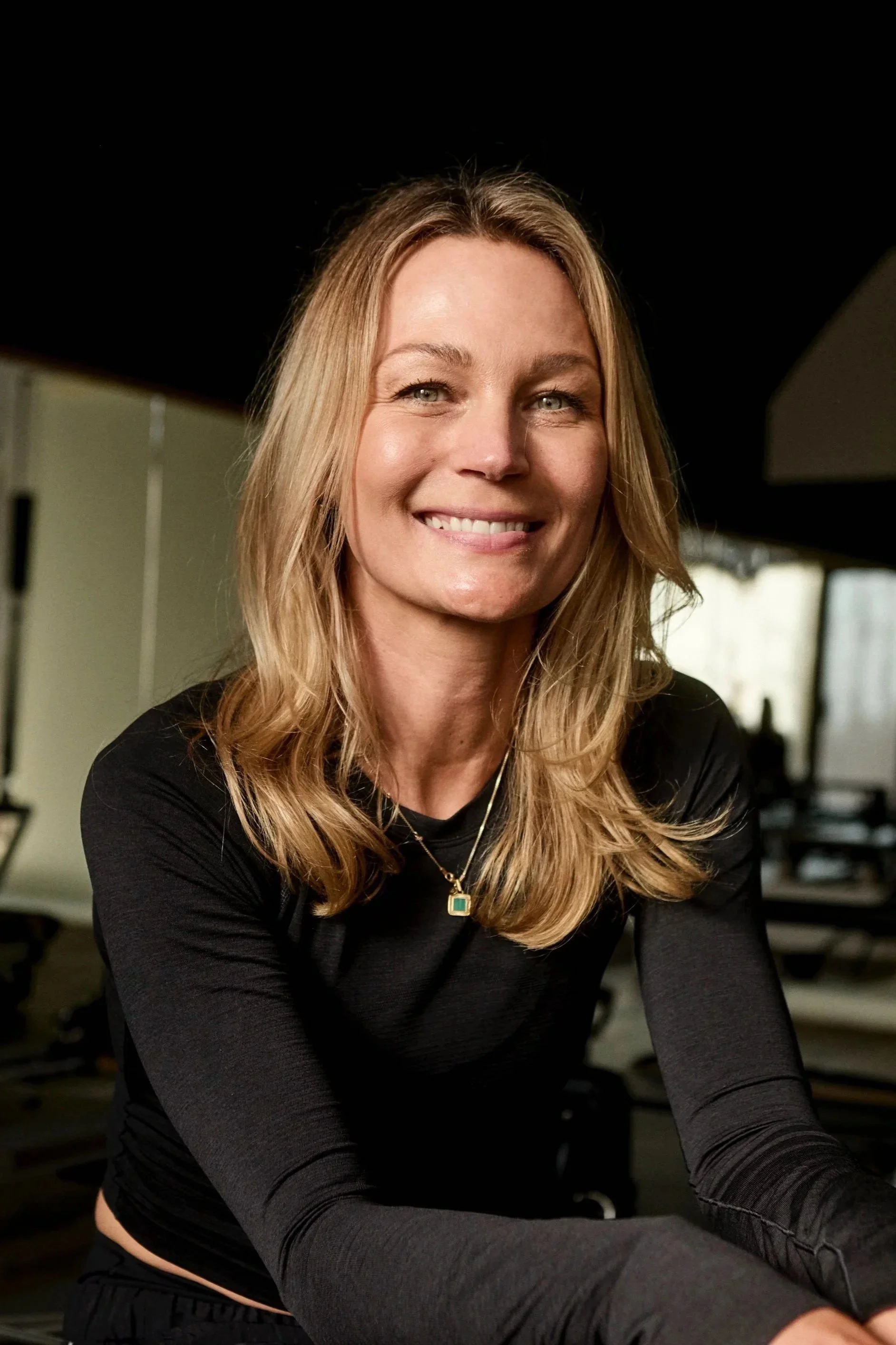 A smiling woman with blonde hair wearing a black long-sleeve shirt and a gold necklace, seated indoors with gym equipment in the background.