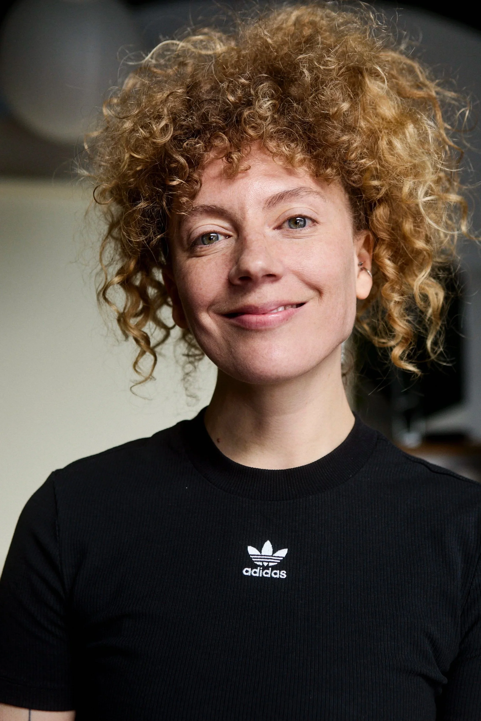 Close-up portrait of a woman with curly red hair, smiling, wearing a black Adidas t-shirt.