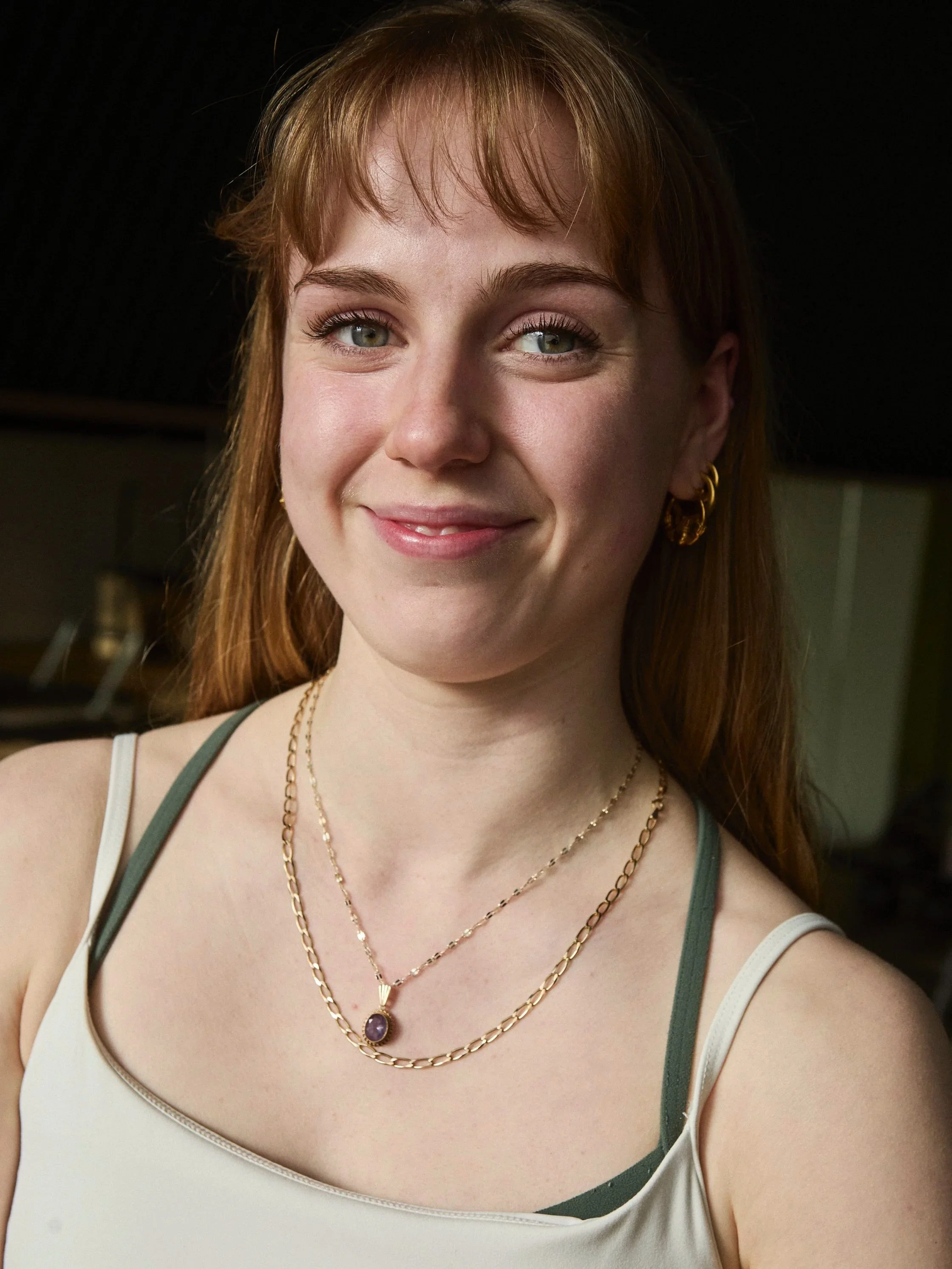 Close-up of a young woman with red hair, blue eyes, wearing gold earrings, layered gold necklaces with pendants, and a sleeveless top with green straps, smiling at the camera.