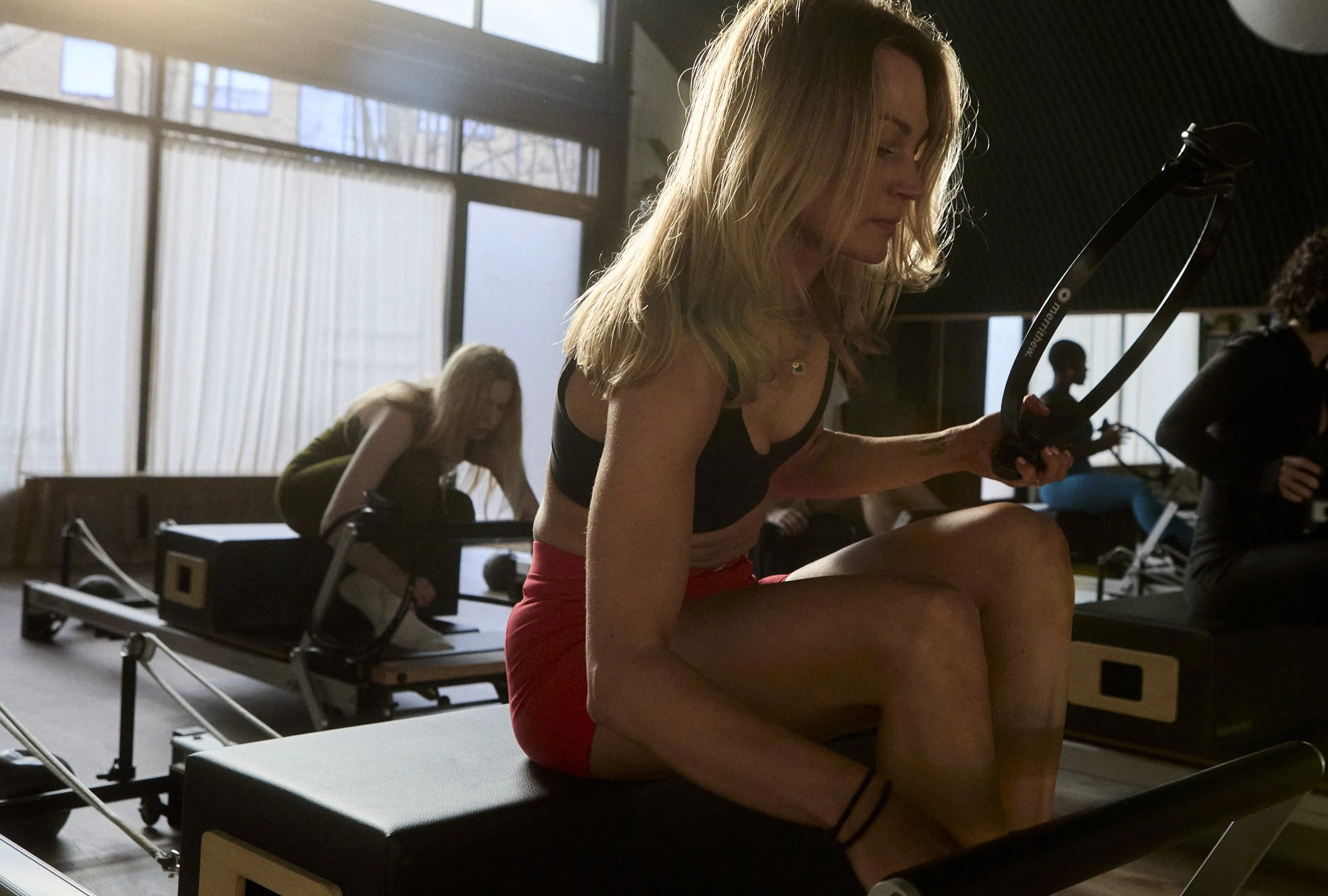 Woman smiling while sitting on a Pilates reformer in a gym studio with other people exercising in the background.