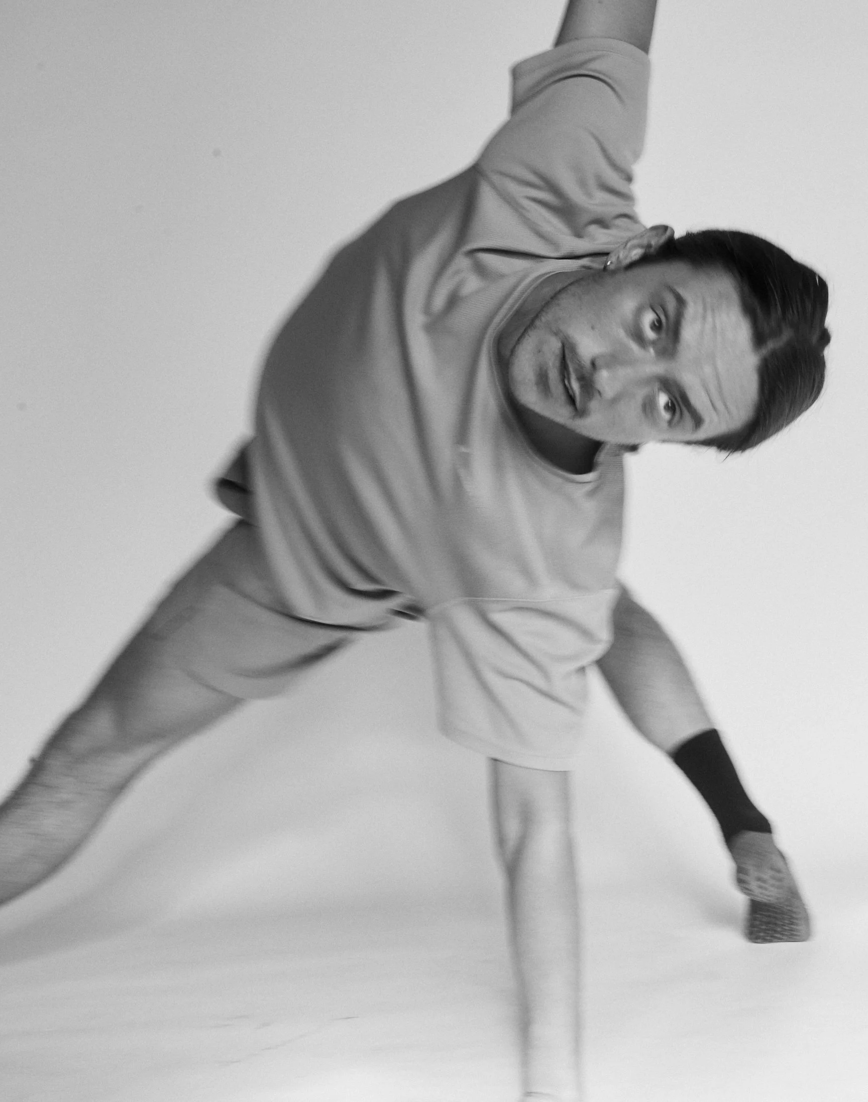 Person sitting on a Pilates reformer with eyes closed, wearing a sweater and holding feet in a stretching pose, indoor setting.