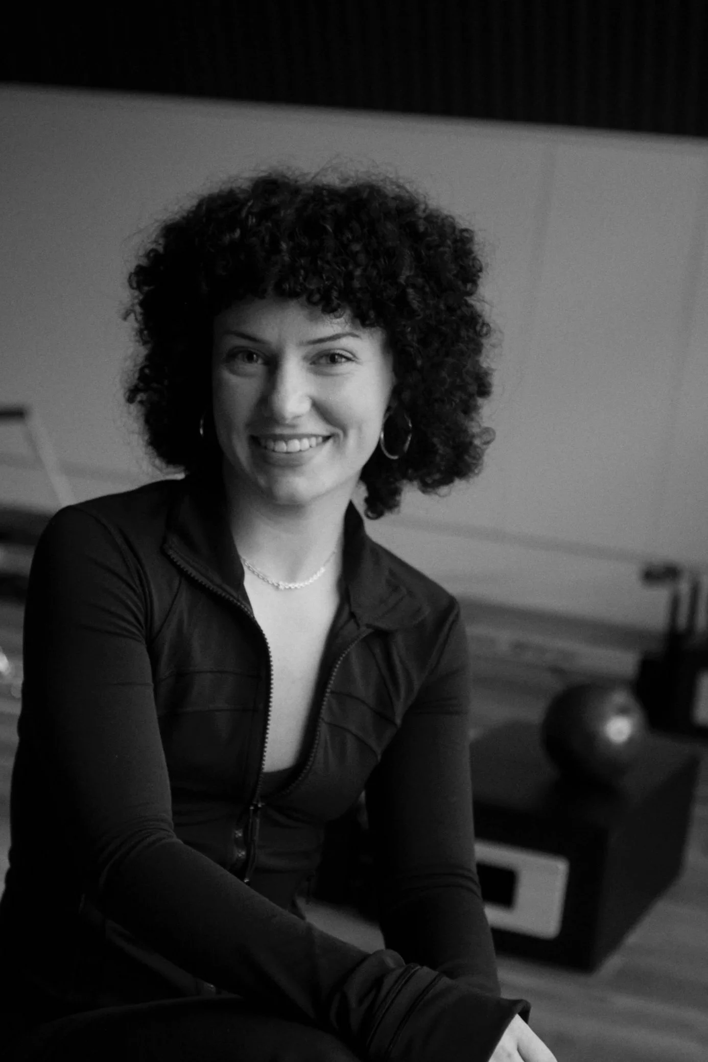Black and white photo of a woman with curly hair smiling, wearing a jacket and hoop earrings, sitting indoors with a blurred background.