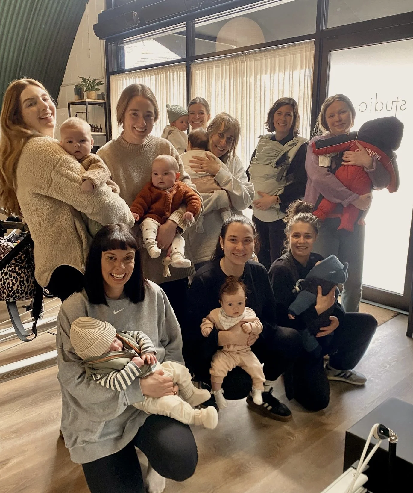 A group of women and children posing together indoors, some holding babies, in front of a glass door with curtains.