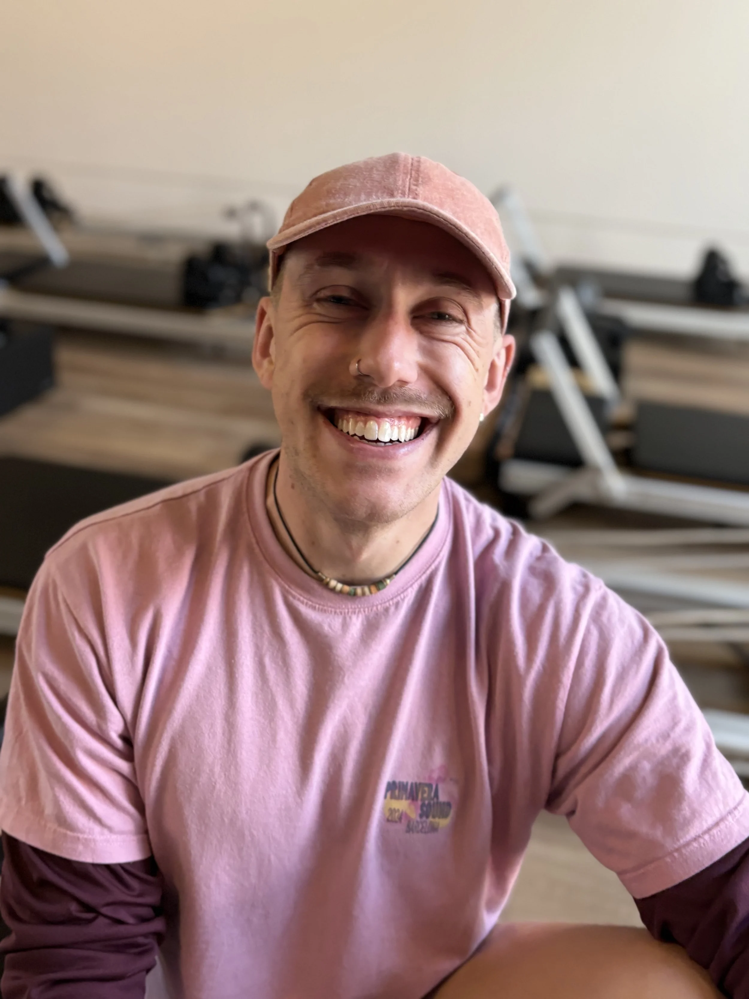 A smiling man wearing a pink cap and pink t-shirt sitting indoors with a blurred background of stacked workout equipment.