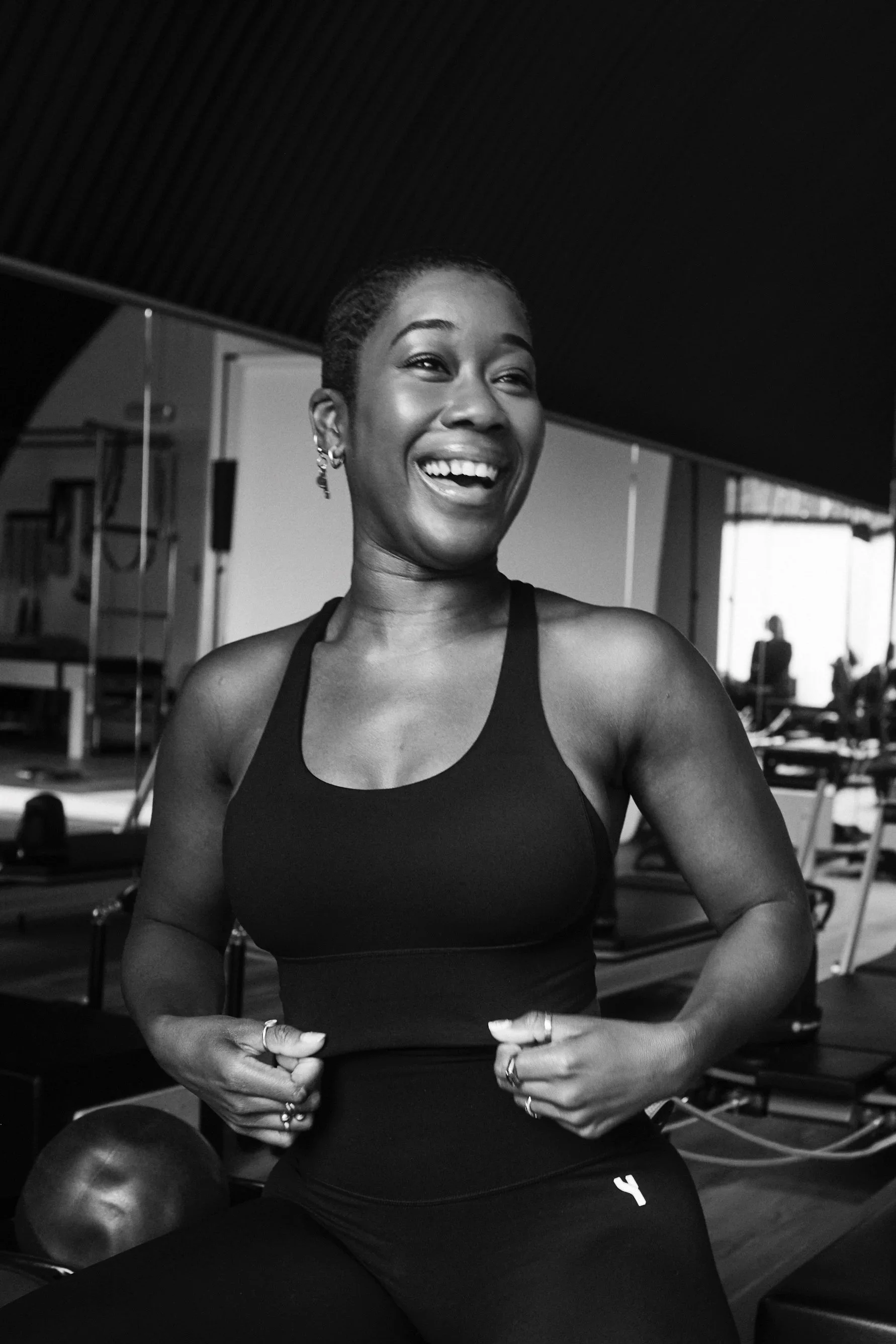A smiling woman in athletic wear adjusting her sports top inside a gym with workout equipment in the background.