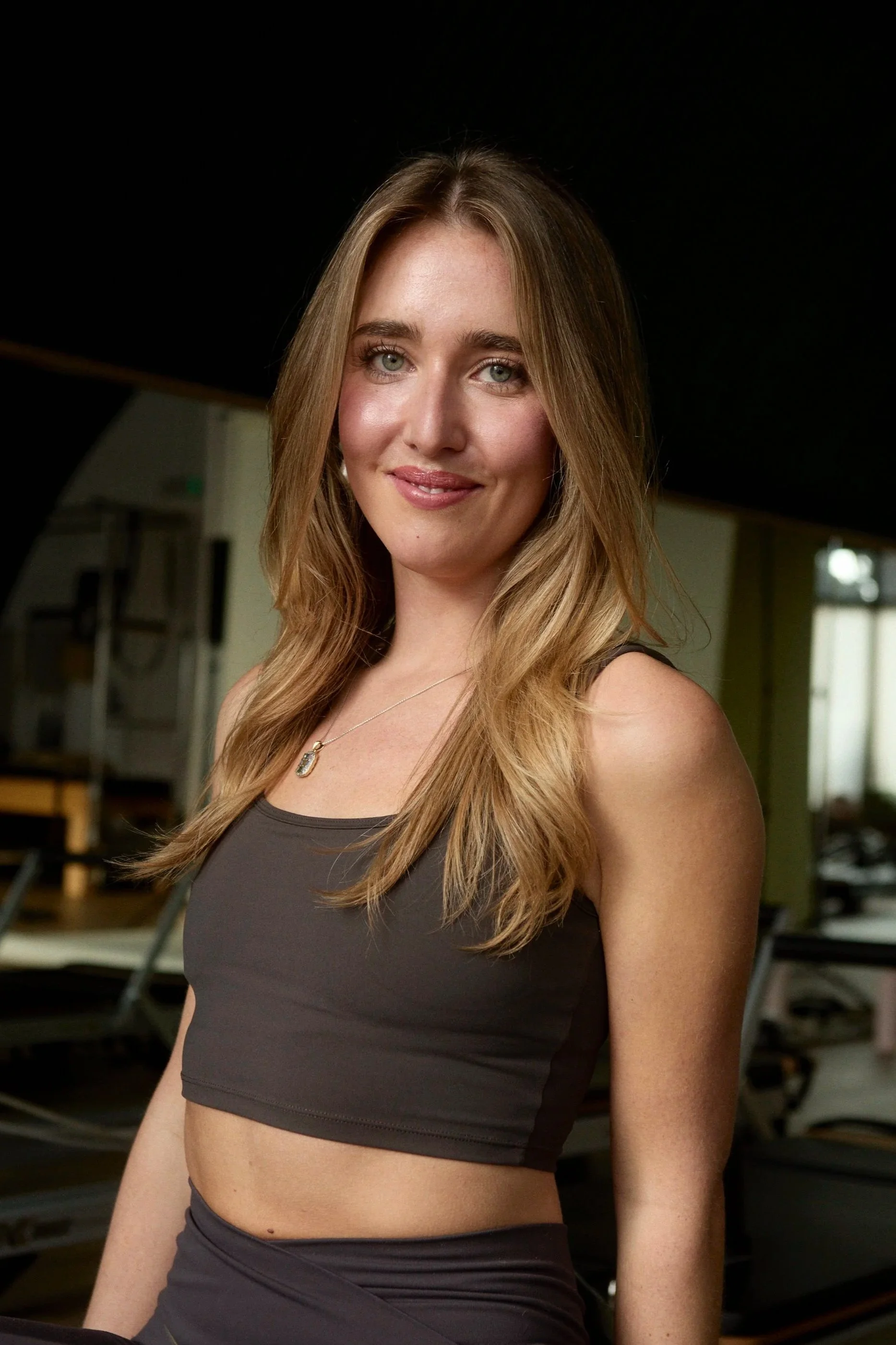A young woman with long blonde hair, light skin, and blue eyes smiling confidently in a gym setting.