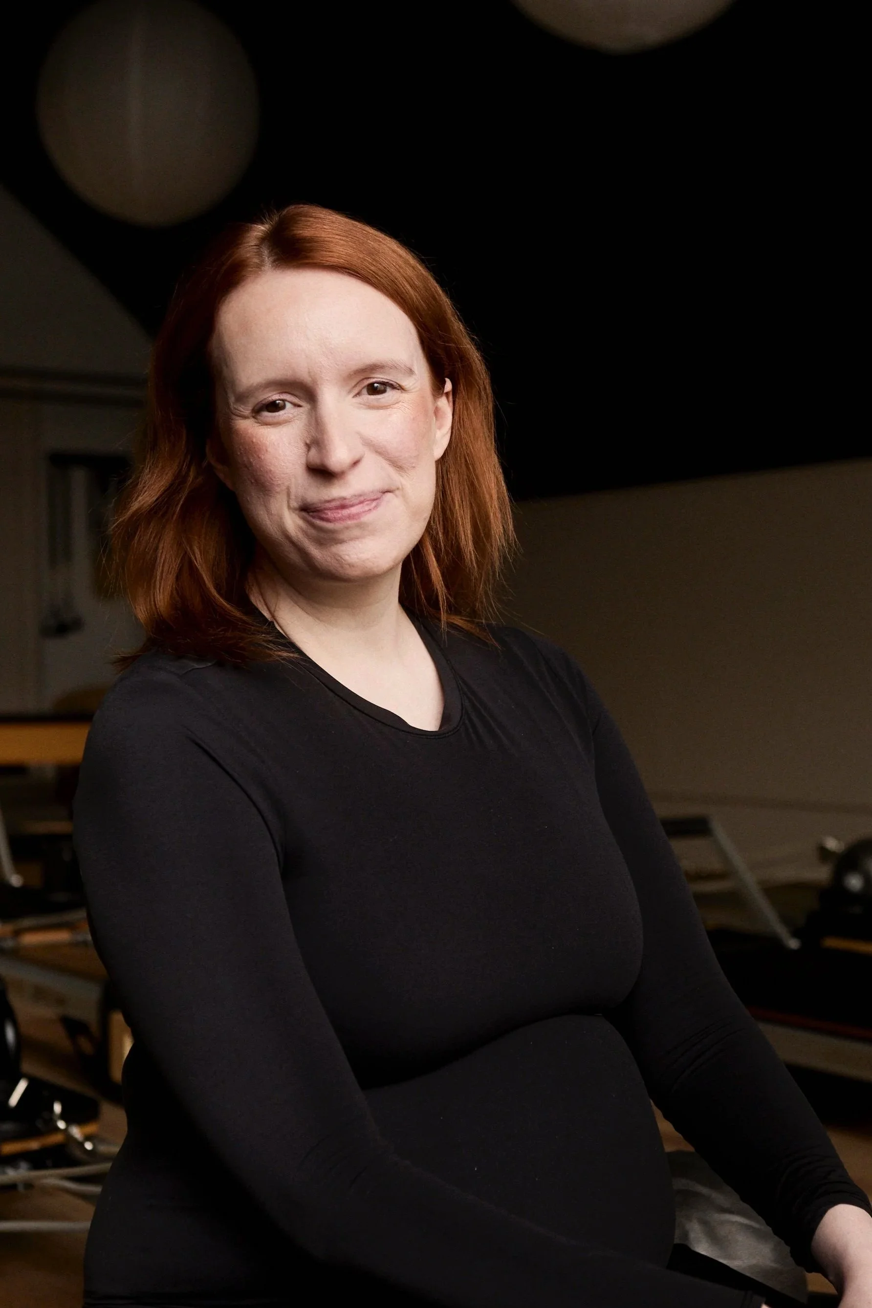 A woman with red hair wearing a black long-sleeve shirt, smiling in a dark room with musical equipment in the background.
