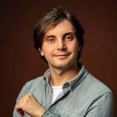 A man with brown hair and a slight smile, wearing a light gray shirt and white T-shirt, posing against a brown background.