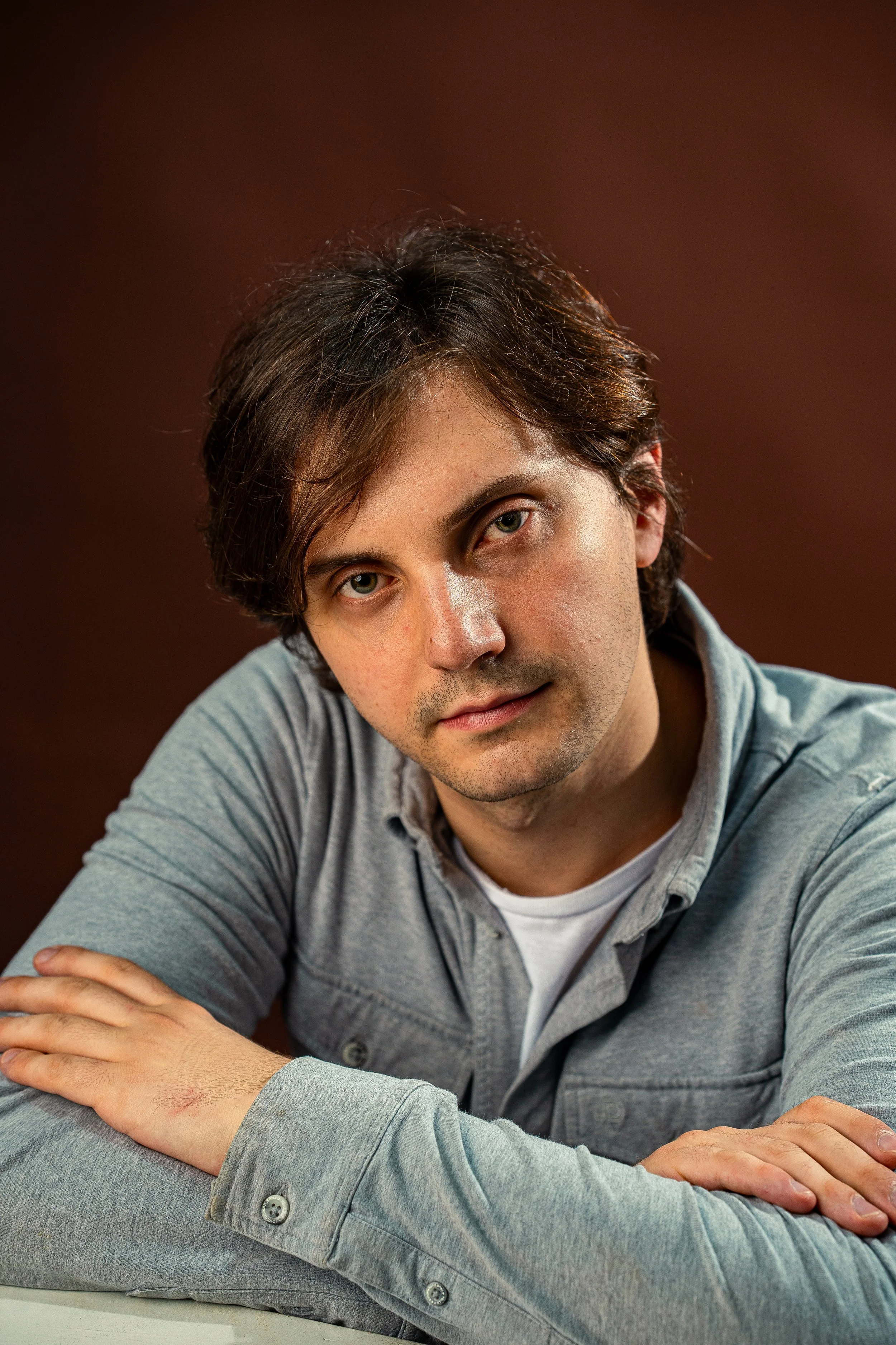 Young man with brown hair and hazel eyes wearing a gray shirt, sitting at a table with his arms crossed, looking at the camera against a dark background.
