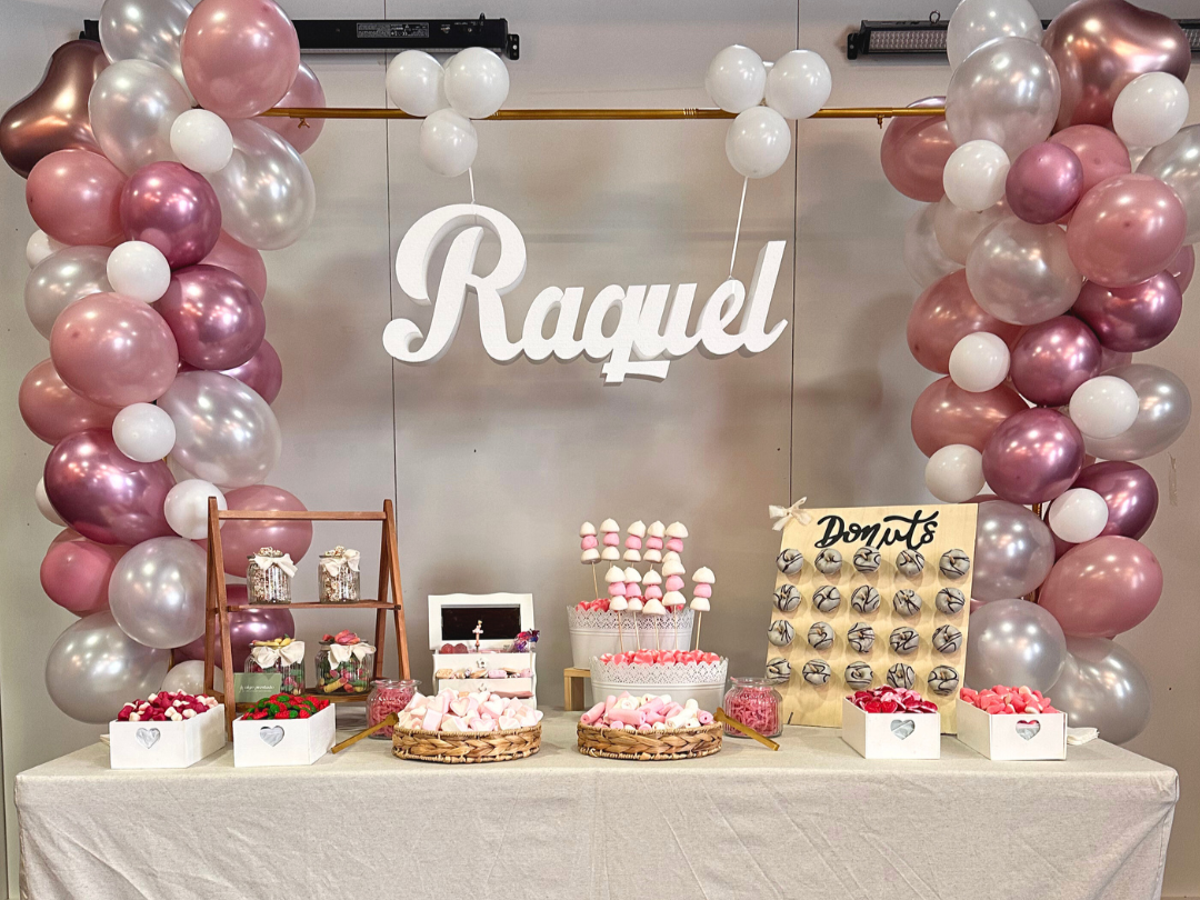 Mesa decorada con globos en tonos rosa, blanco y gris, con un cartel que dice "Raquel" y una mesa con dulces como cakepops, donas y chocolates para una celebración.