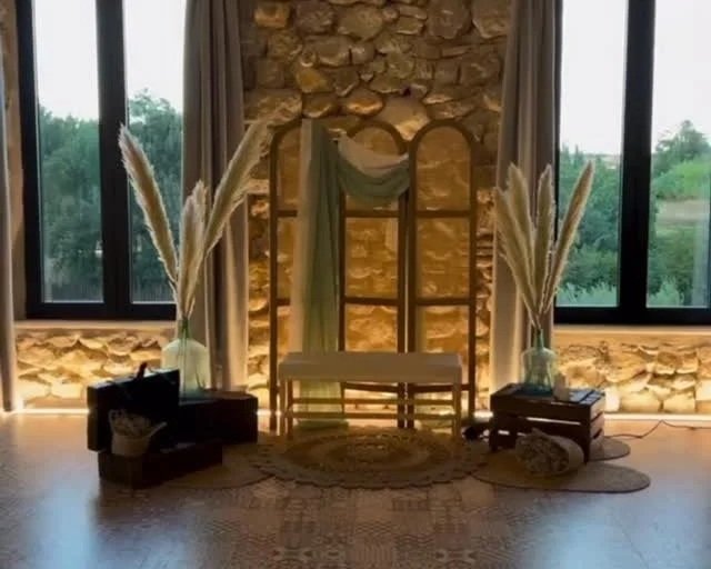 Decoración de interior con una silla en el centro, cortinas, y plantas de pampa en jarrones, con paredes de piedra y ventanas que muestran un paisaje natural.