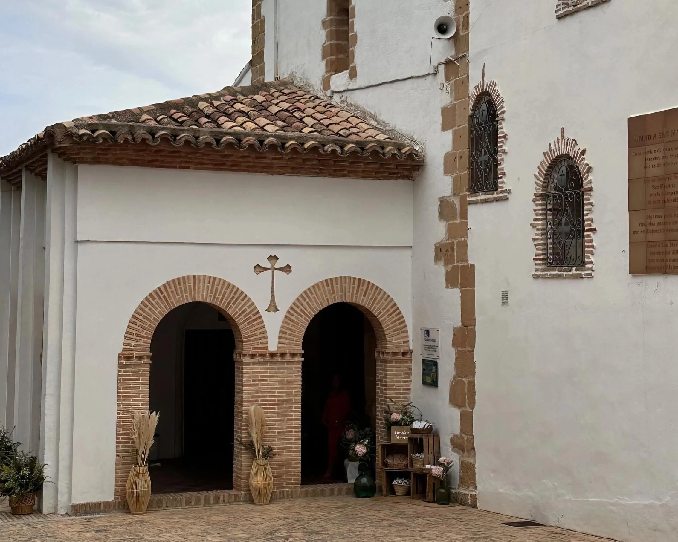 Fachada de una iglesia con paredes blancas, ventanas con rejas de herrería, una cruz y decoración de palmas en la entrada.