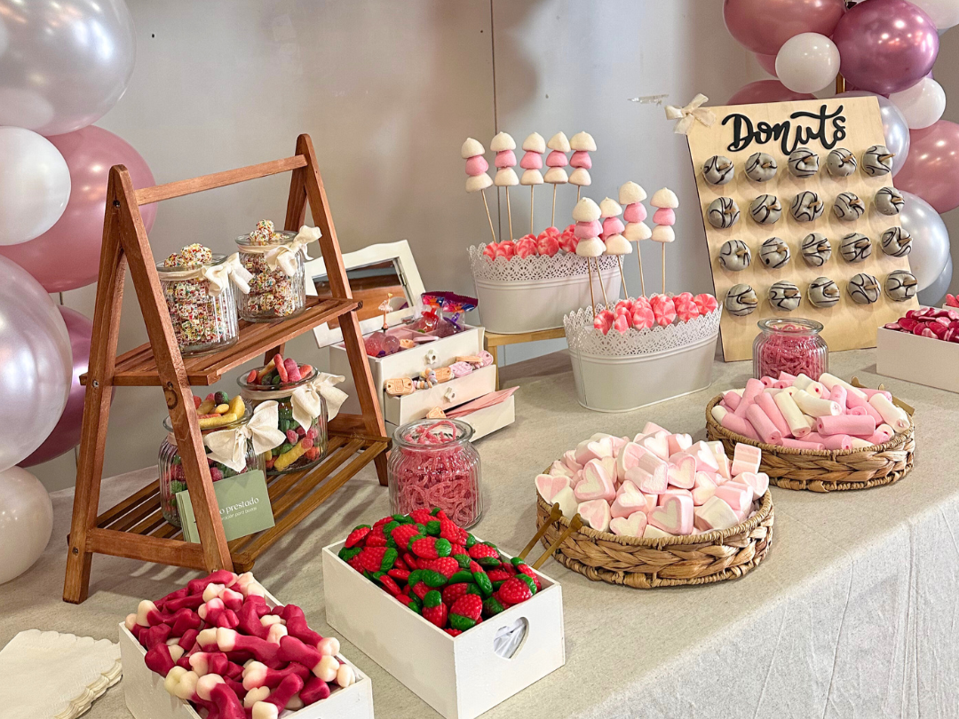 Mesa decorada con dulces y golosinas, incluyendo piruletas, caramelos, y chocolates, con guirnaldas de globos de colores pastel en el fondo, y un cartel que dice "Donas".