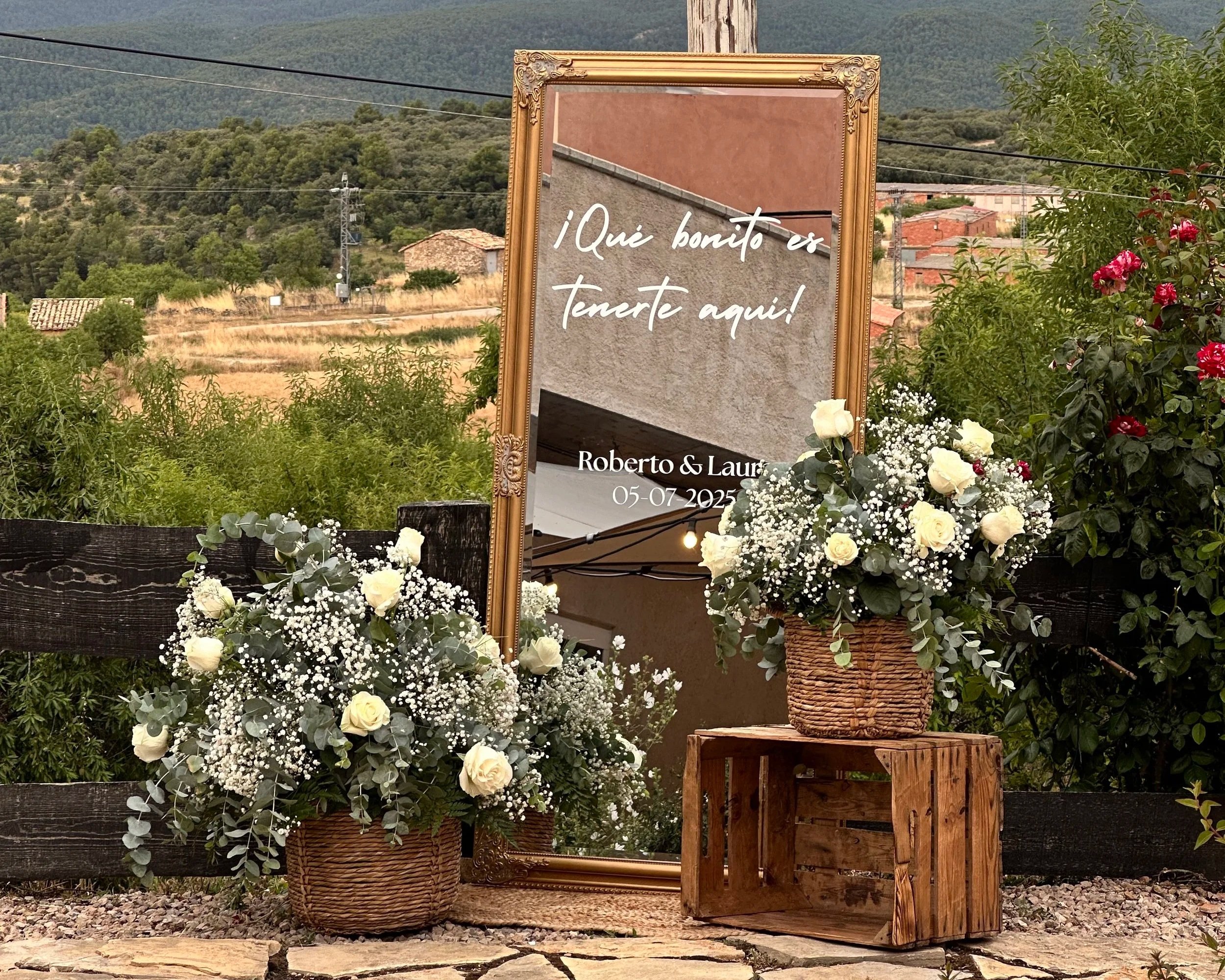 Cartel decorativo con la frase "¡Qué bonito es tenerte aquí!" acompañado de flores blancas en cestas y un fondo de naturaleza y montañas.