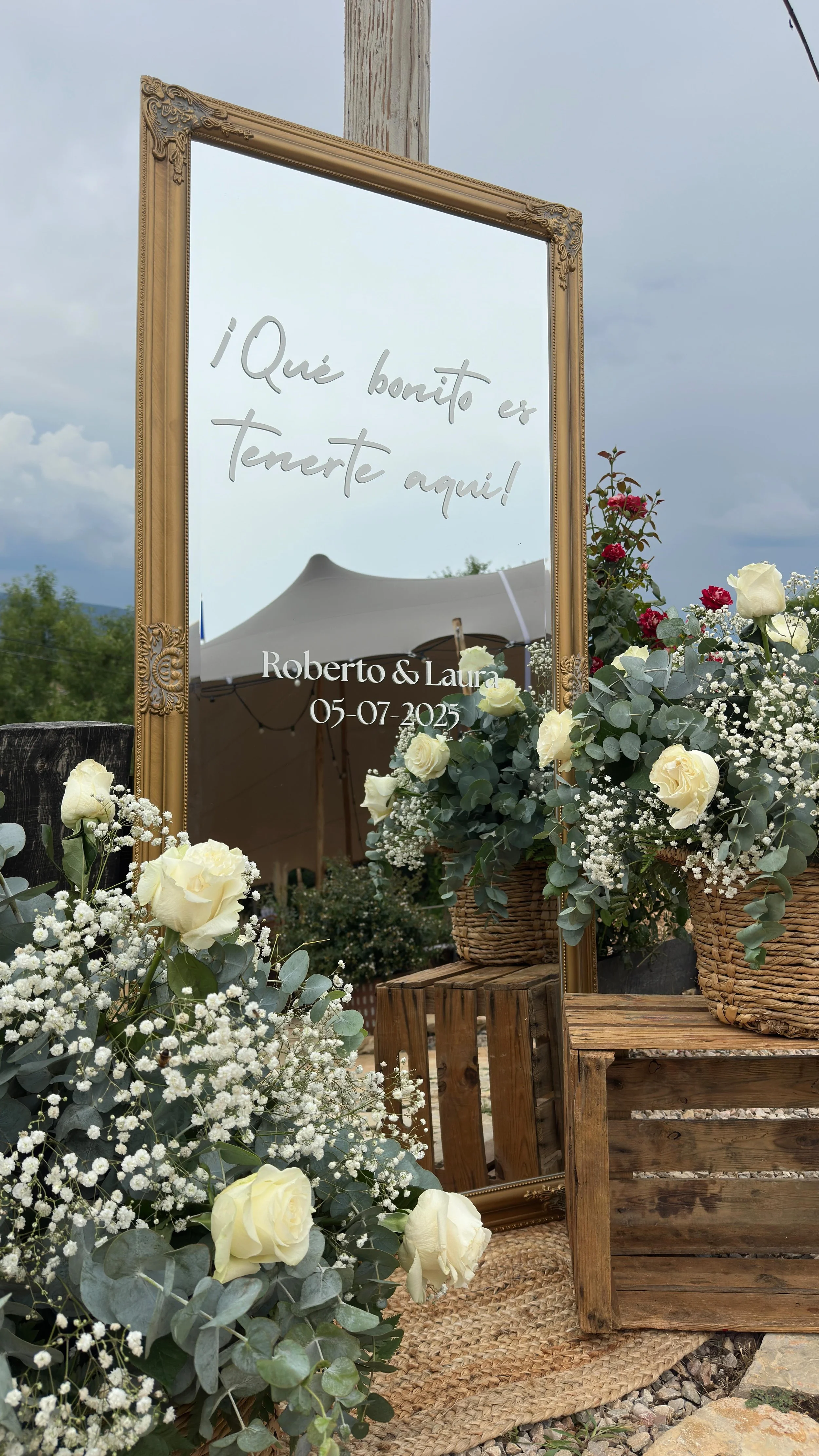 Espejo decorativo con mensaje en español que dice '¡Qué bonito es tenerte aquí!' junto a flores blancas en un entorno de celebración al aire libre.