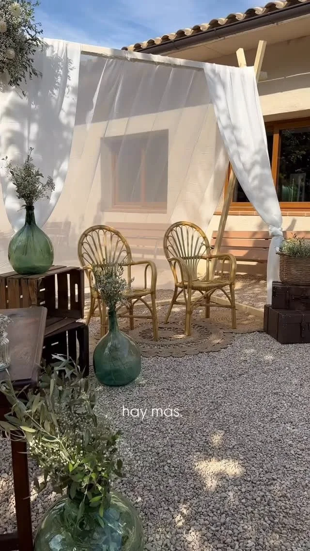 Zona de descanso con sillas de mimbre, macetas de vidrio con plantas, cortina de tela blanca y un cobertizo exterior en un patio de piedra.