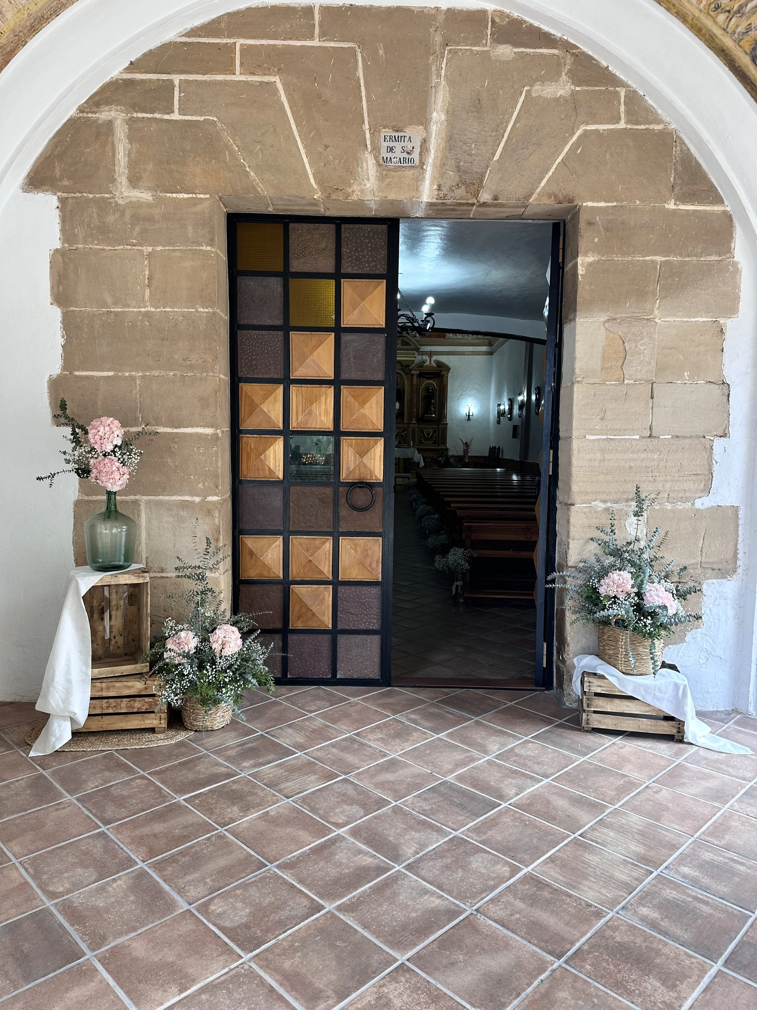 Entrada a una iglesia con una puerta de madera con marco de hierro, decorada con arreglos de flores en cestas de mimbre y tablas de madera. Hay un letrero que dice 'ERMITA DE SAN MÁXARIO' y el interior visible desde la entrada muestra bancos de mader