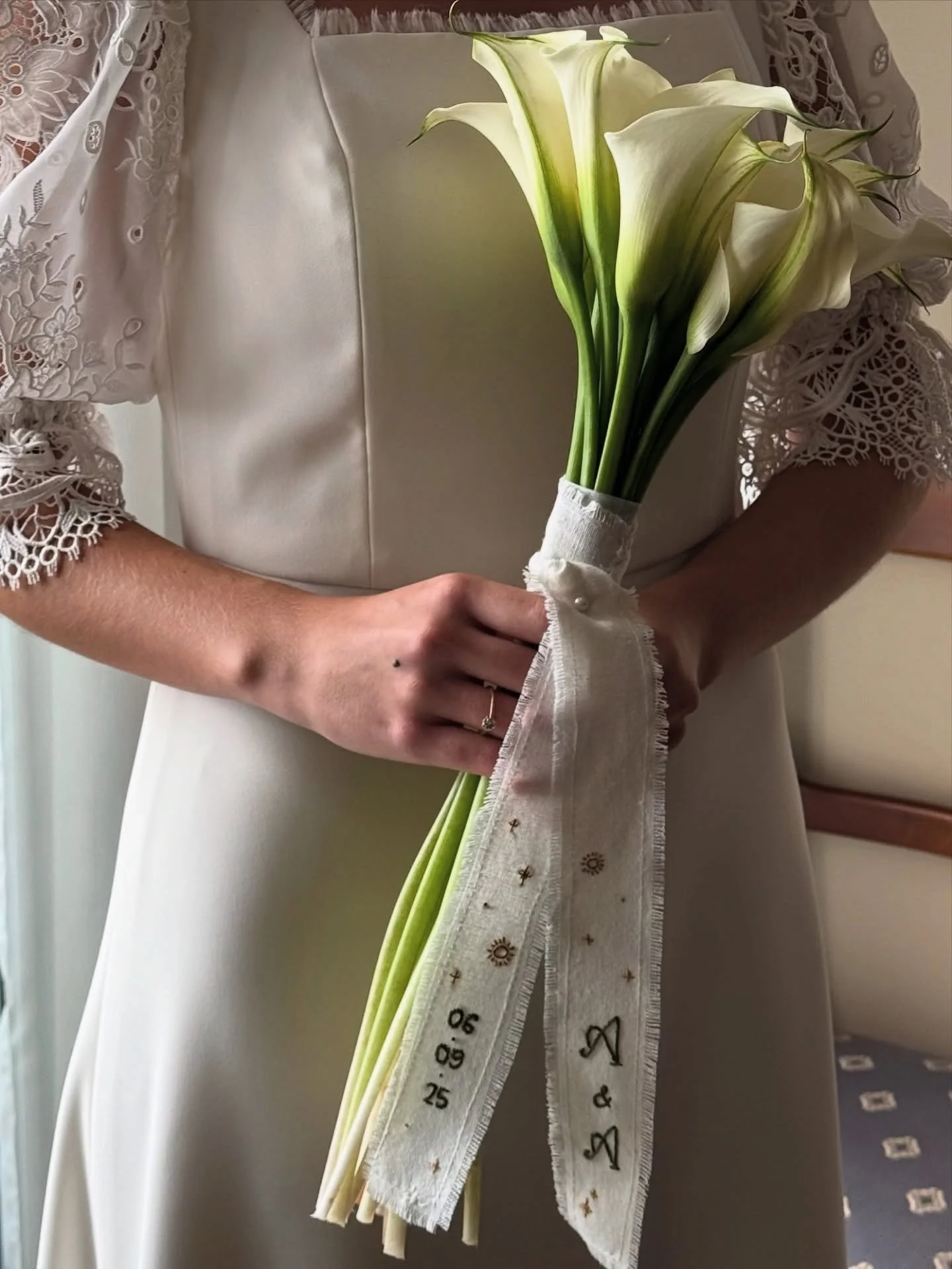 Una mujer sostiene un ramo de flores de lirios blancos con tallos largos, atados con una cinta decorativa blanca que tiene bordados dorados, en un vestido blanco con encaje en las mangas.