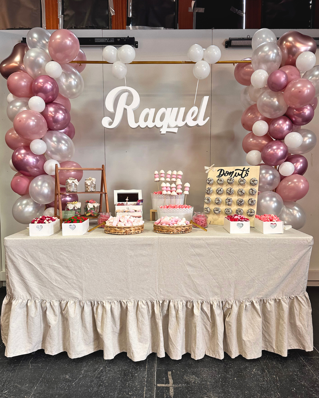 Mesa decorada con globos en tonos rosa y blanco, con un letrero que dice 'Raquel' y un puesto de dulces con donas y malvaviscos, decoraciones para fiesta.