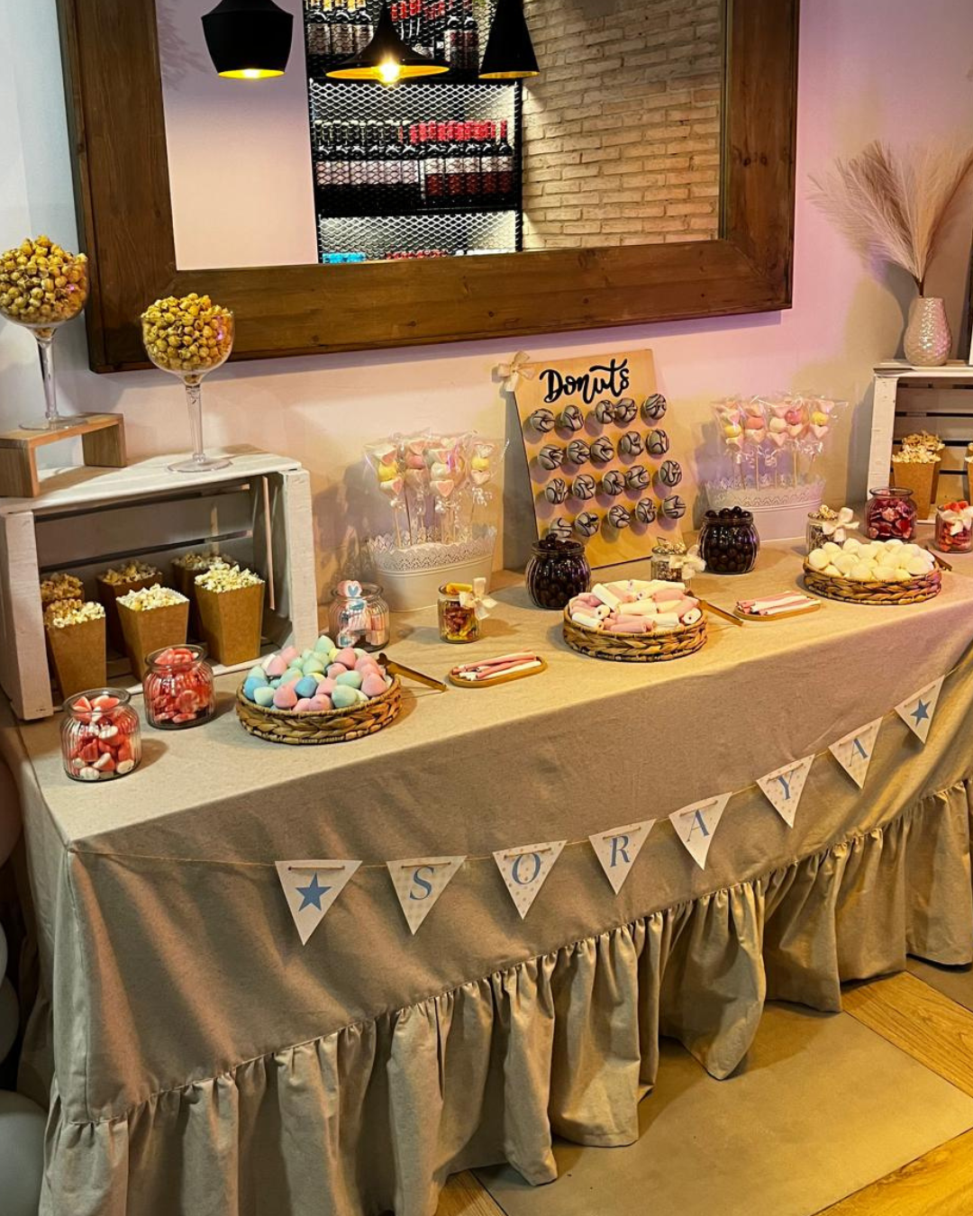 Mesa decorada con dulces y donas para una celebración, con un cartel que dice 'SORRY' y una bandera que dice 'SORAYA', con flores y un espejo en el fondo.