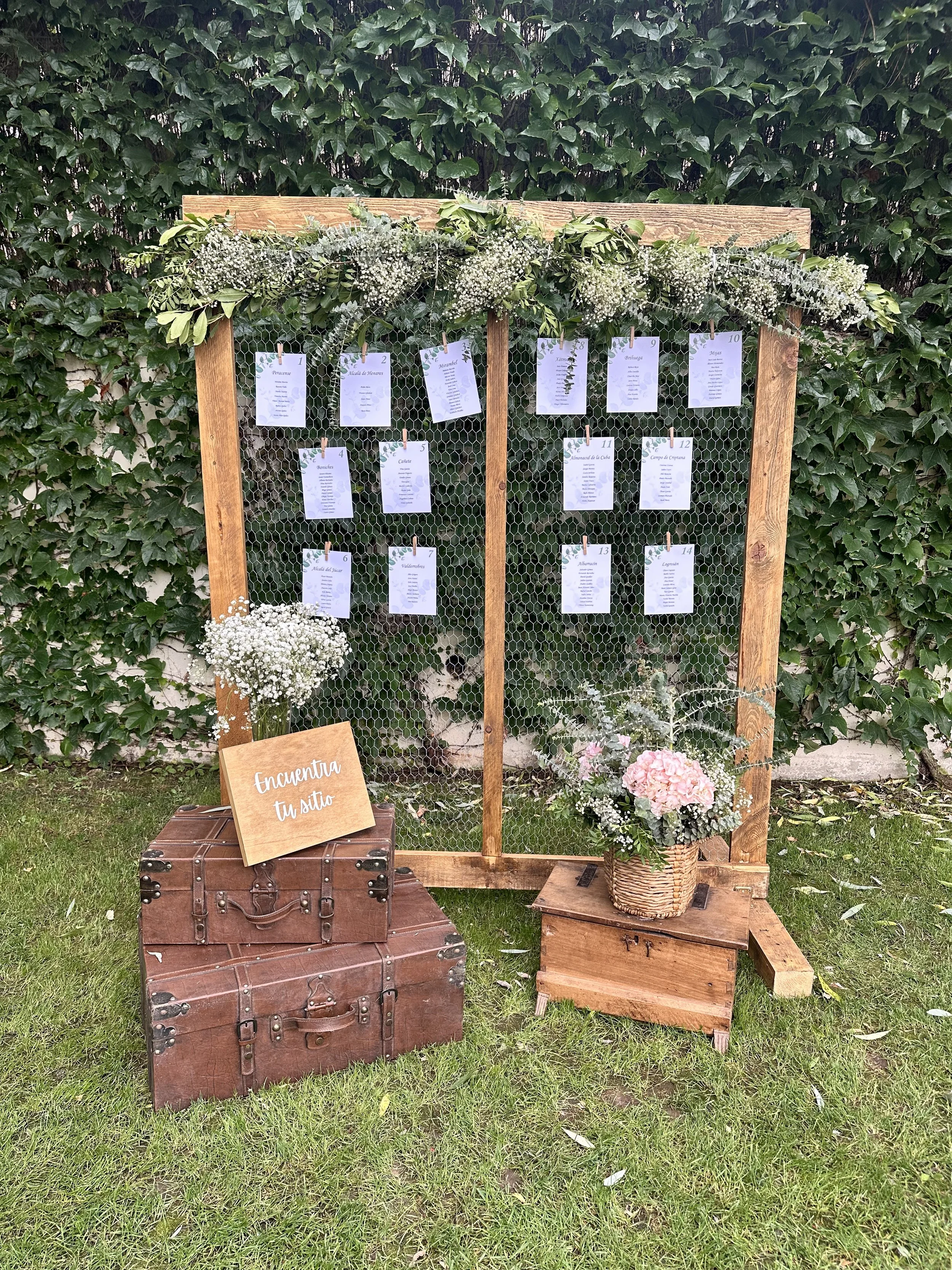 Cartelera de asientos para una boda o evento, decorada con flores blancas y verdes, con rótulo que dice 'Encuentra tu sitio', en un jardín con muro de hiedra.