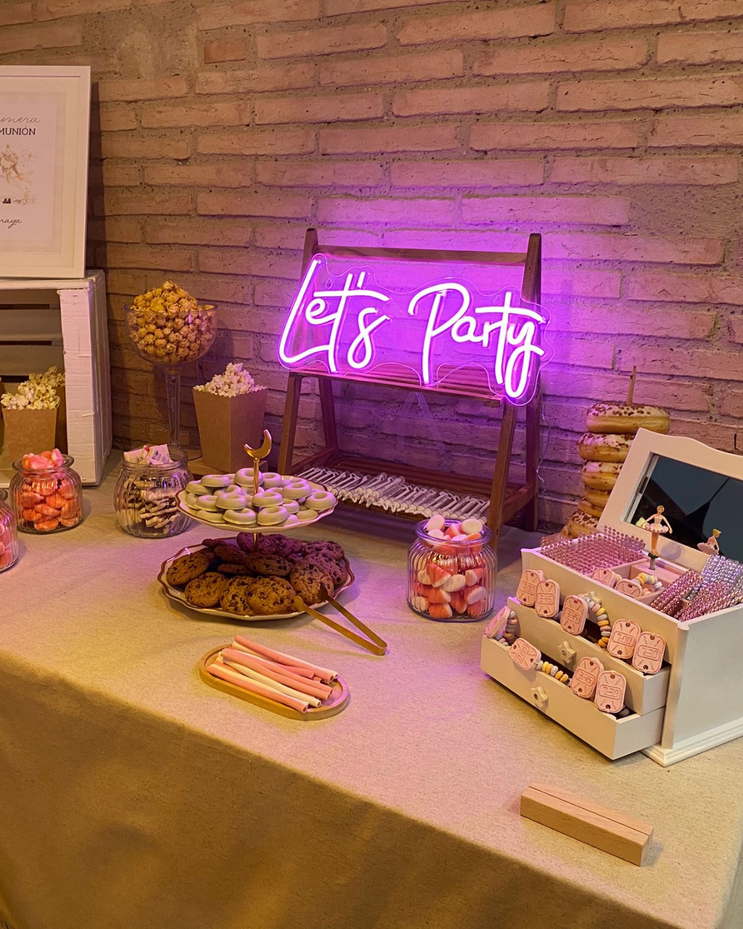 Mesa con dulces y galletas decoradas, decoraciones de fiesta, letrero de neón que dice 'Let's Party', y una caja con accesorios para decorar.