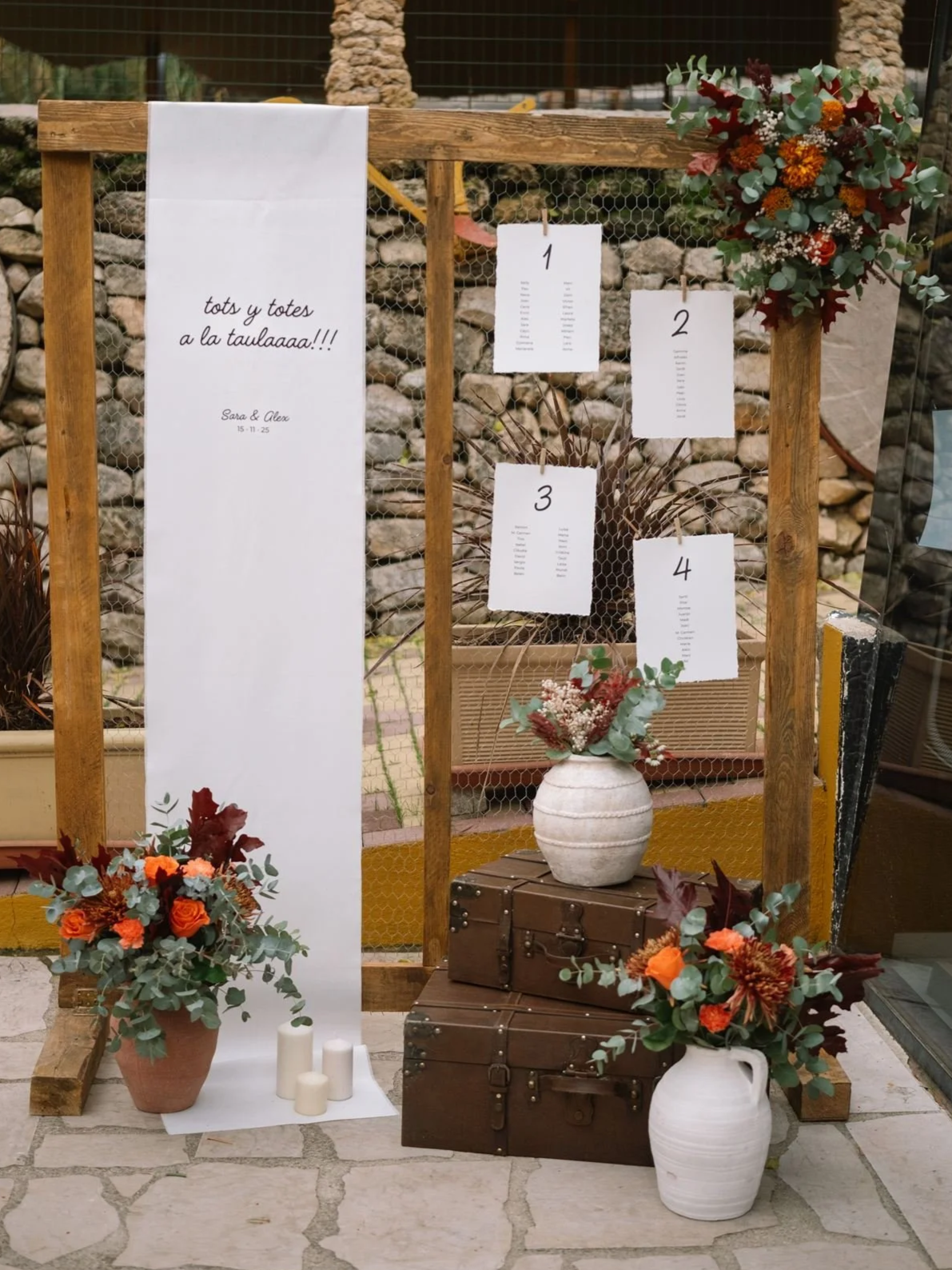 Se observa una decoración con arreglos florales en jarrones, una estructura de madera con malla de gallinero, varias hojas en papel con números y una lista, y velas blancas en el suelo, probablemente para una boda o evento especial.