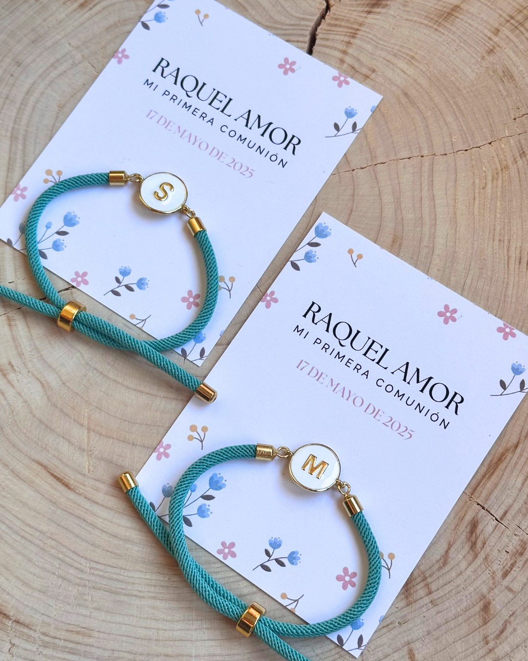 Dos pulseras de cuerda azul con tapas blancas y letras doradas, colocadas sobre invitaciones de mariposas, flores y ramas, que dicen 'Raquel Amor, mi primera comunión' con fechas 17 de mayo de 2023 y 17 de mayo de 2025, en una mesa de madera.