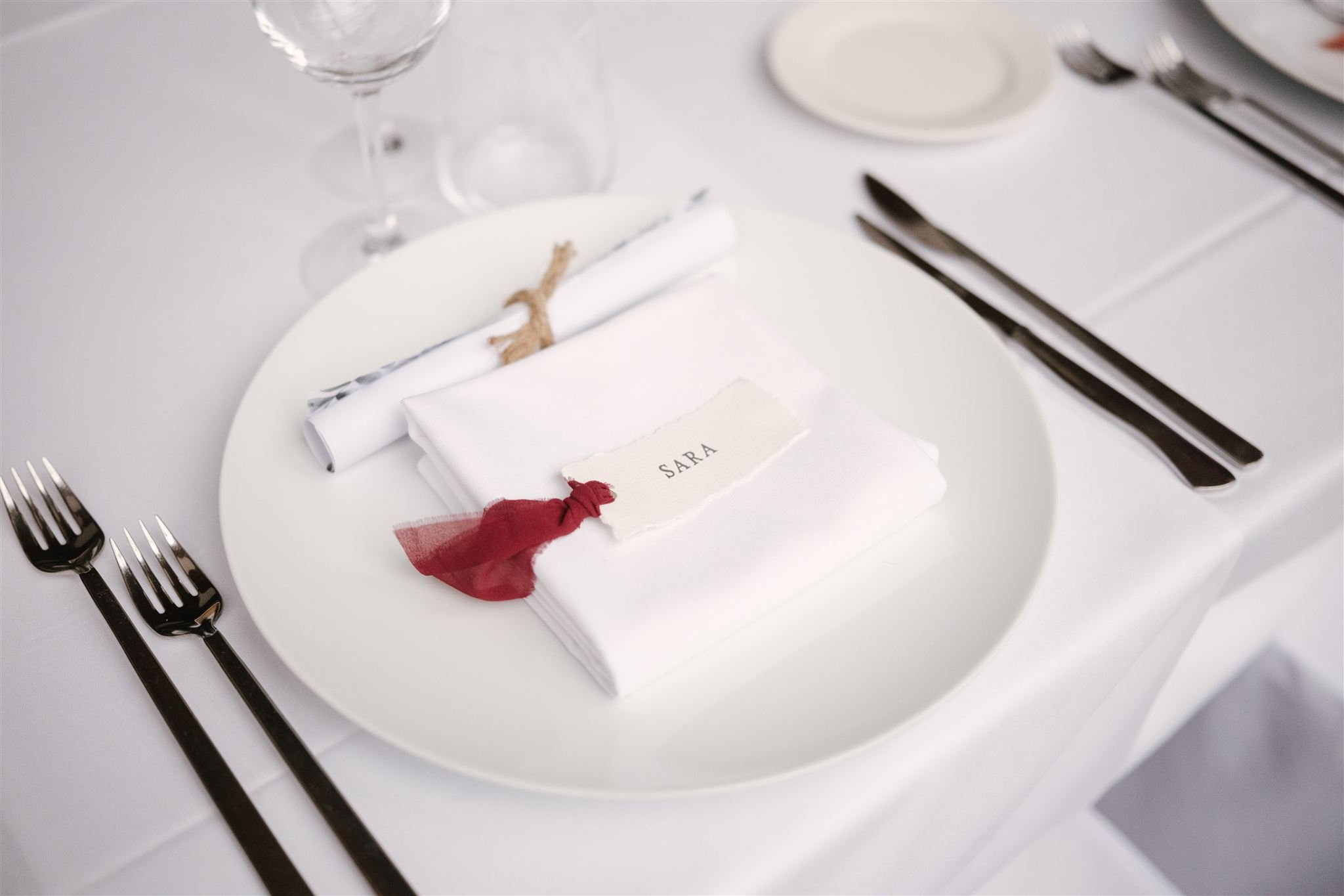 Detalle de un lugar en una mesa con un plato blanco, configurado para una cena formal, incluyendo cubiertos, copas y un servilleta con una etiqueta que dice 'Sara'.