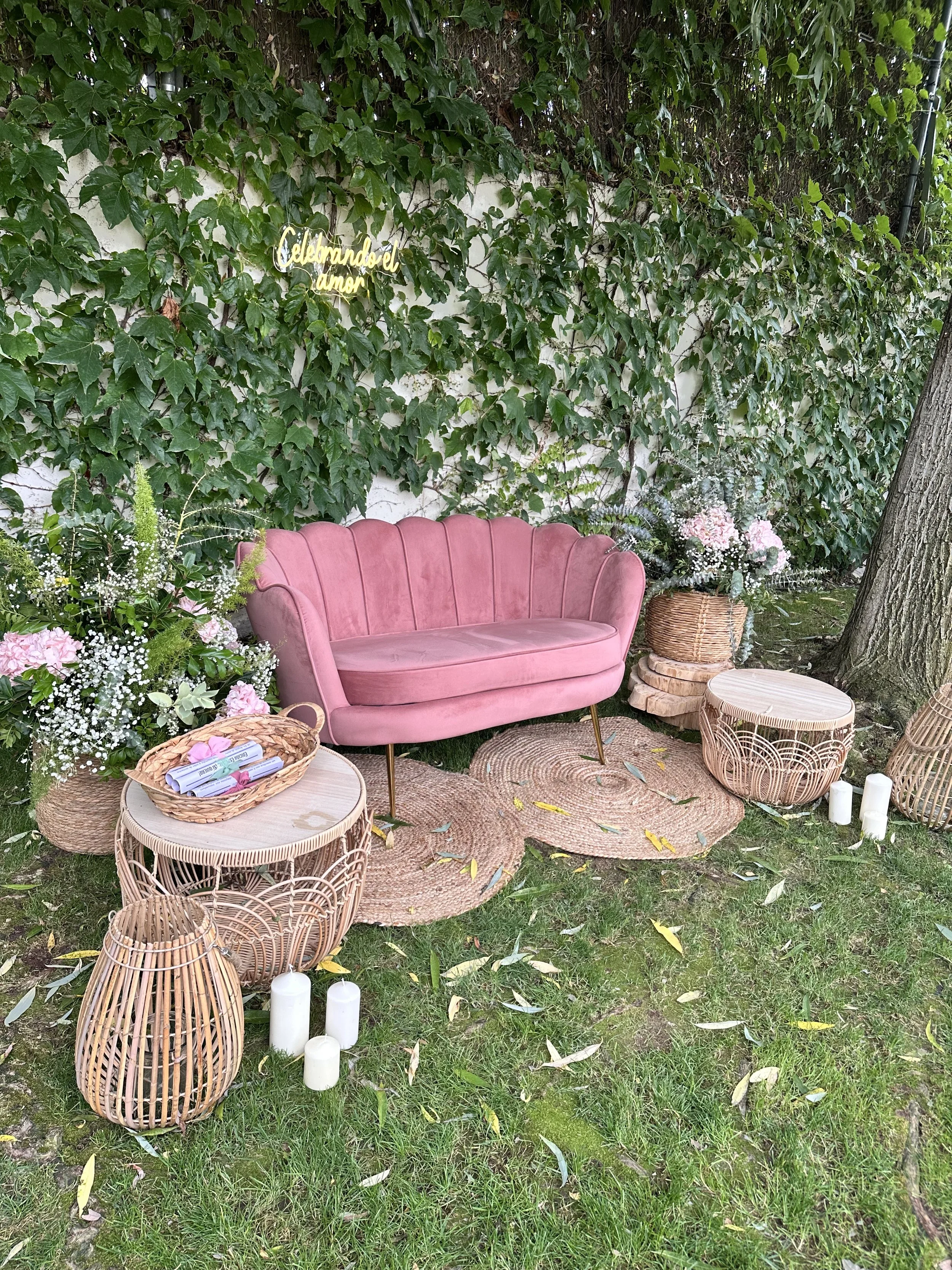 Sofá rosa en un espacio al aire libre con plantas y decoración, incluyendo cestas de mimbre, velas y un letrero que dice 'Celebrando el amor'