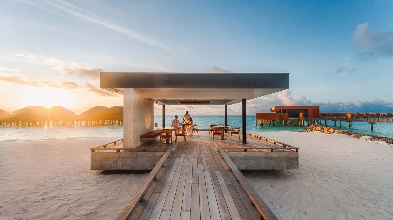 Utomhusrestaurang vid havet med solnedgång, sandstrand och havsutsikt, bord och stolar under ett modernt tak.