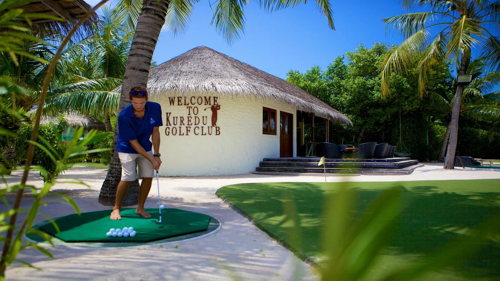 En person spelar golf på en putting green framför Kuredu Golf Club, omgiven av palmer.