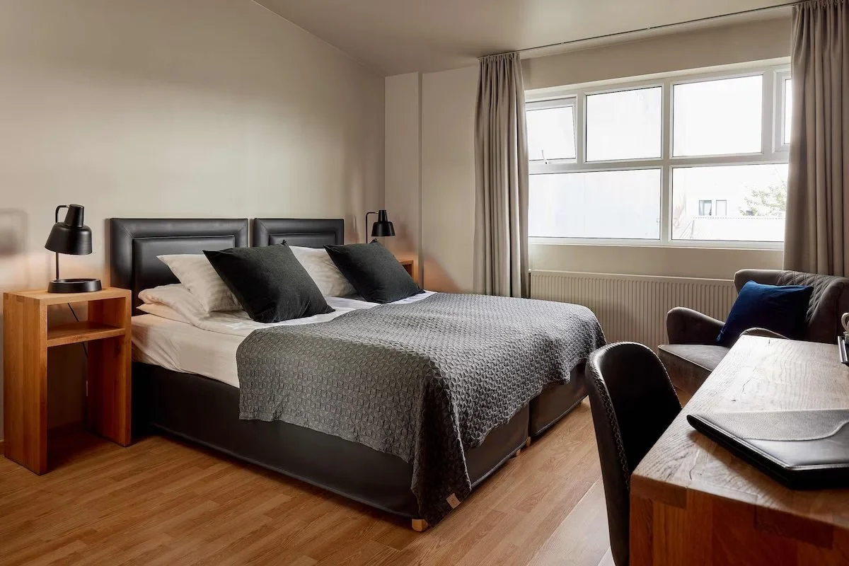Ett smakfullt inrett hotellrum på hotel FRON, Reykjavik med dubbelsäng, två svarta sängbord med lampor, stor fönstervägg med gardiner, ett skrivbord med medlemsmaterial och en bekväm stol. Ett rymligt och ljust rum.
