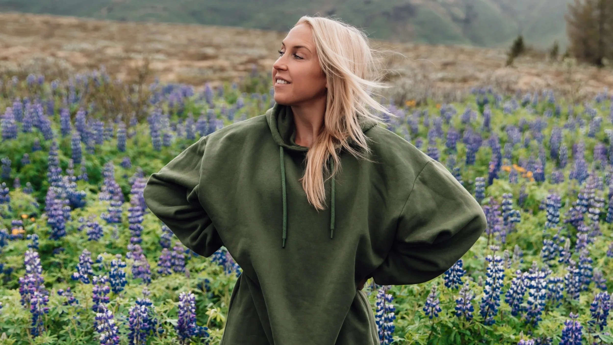 Elin Härkönen i en grön hoodie står i ett fält med blåa lupiner. Hon tittar åt sidan och ler medan vinden rör vid hennes hår.