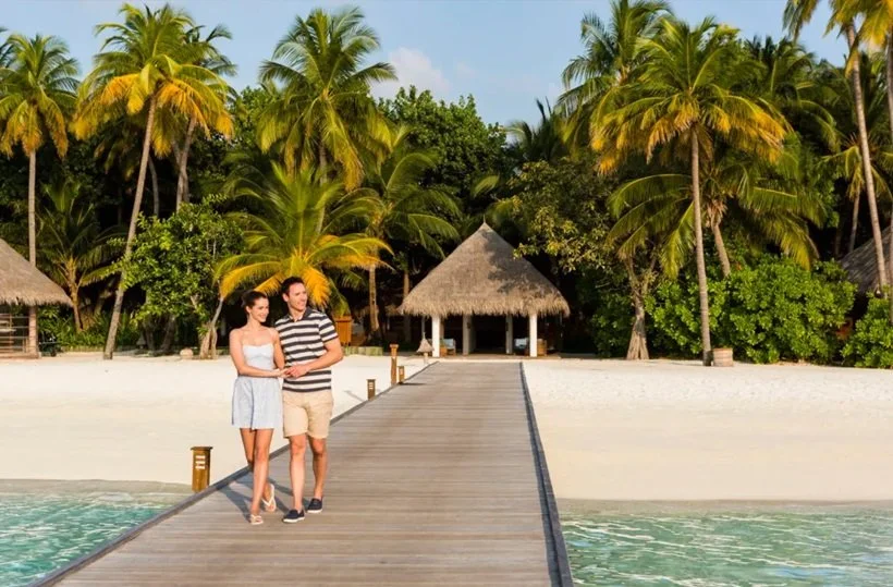 Par går på en träbrygga vid en tropisk strand med palmer och en hydda i bakgrunden.