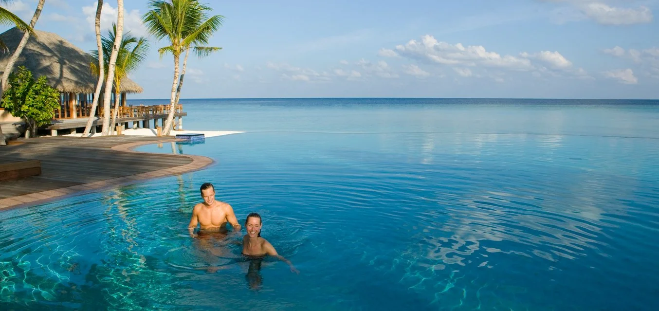 Två personer i en pool vid en tropisk strand, palmer och en bungalow.