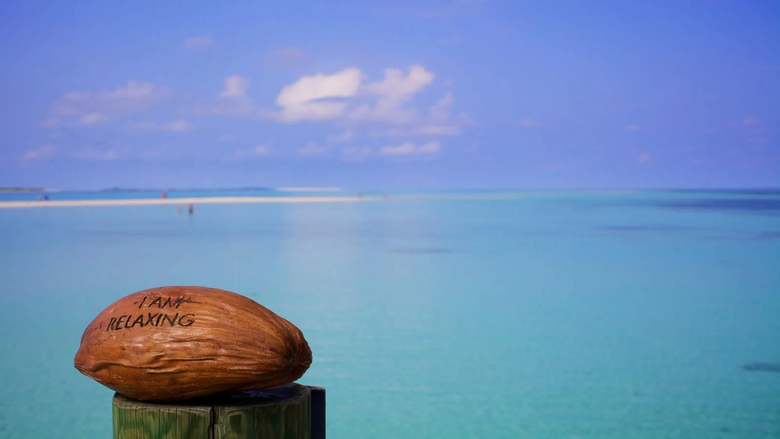 Kokosnöt med texten "I am relaxing" på en stolpe vid en tropisk strand med klart blått vatten och himmel.