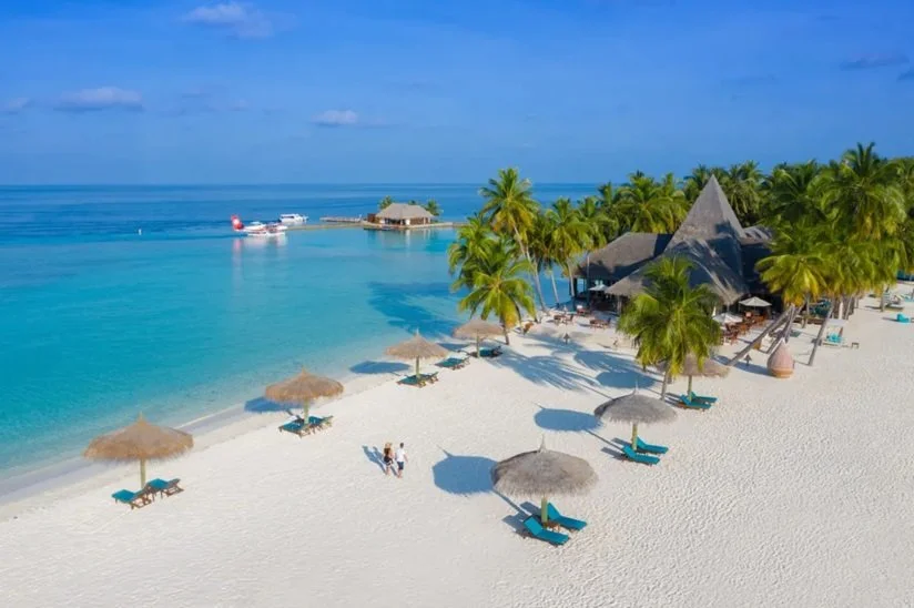 En tropisk strand med vit sand, palmer och parasoller. Turkosblått hav, en bungalow vid vattnet och en sjöflygplan i bakgrunden. Två personer promenerar på stranden.