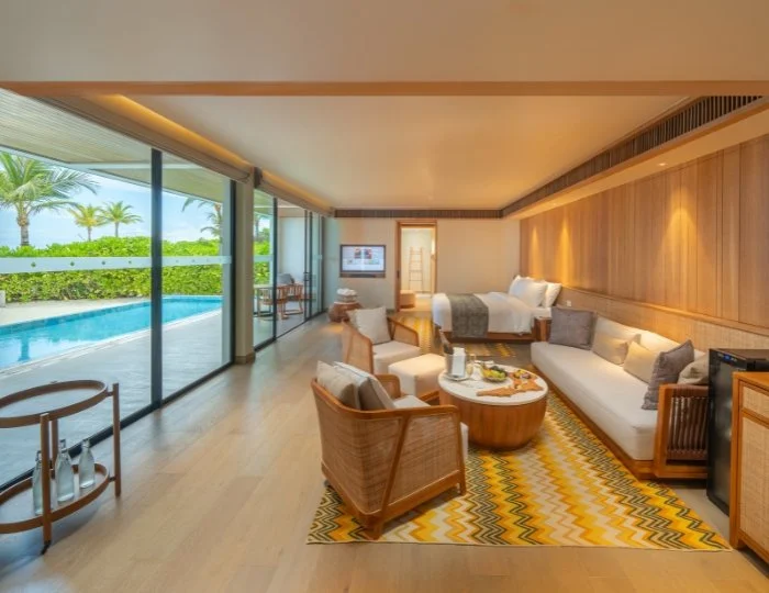 Lyxigt hotellrum med träinredning, sittgrupp och öppen planlösning. Stora fönster vetter mot en pool och grönska utanför. Färgglada kuddar och matta pryder rummet.