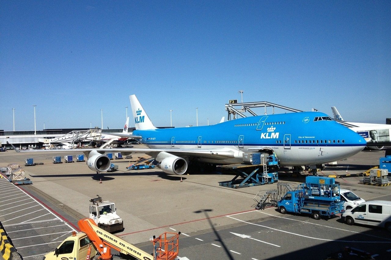 Flygplan från KLM står parkerat vid en gate på en flygplats. Flera fordon och bagagevagnar syns på marken. En klarblå himmel i bakgrunden.