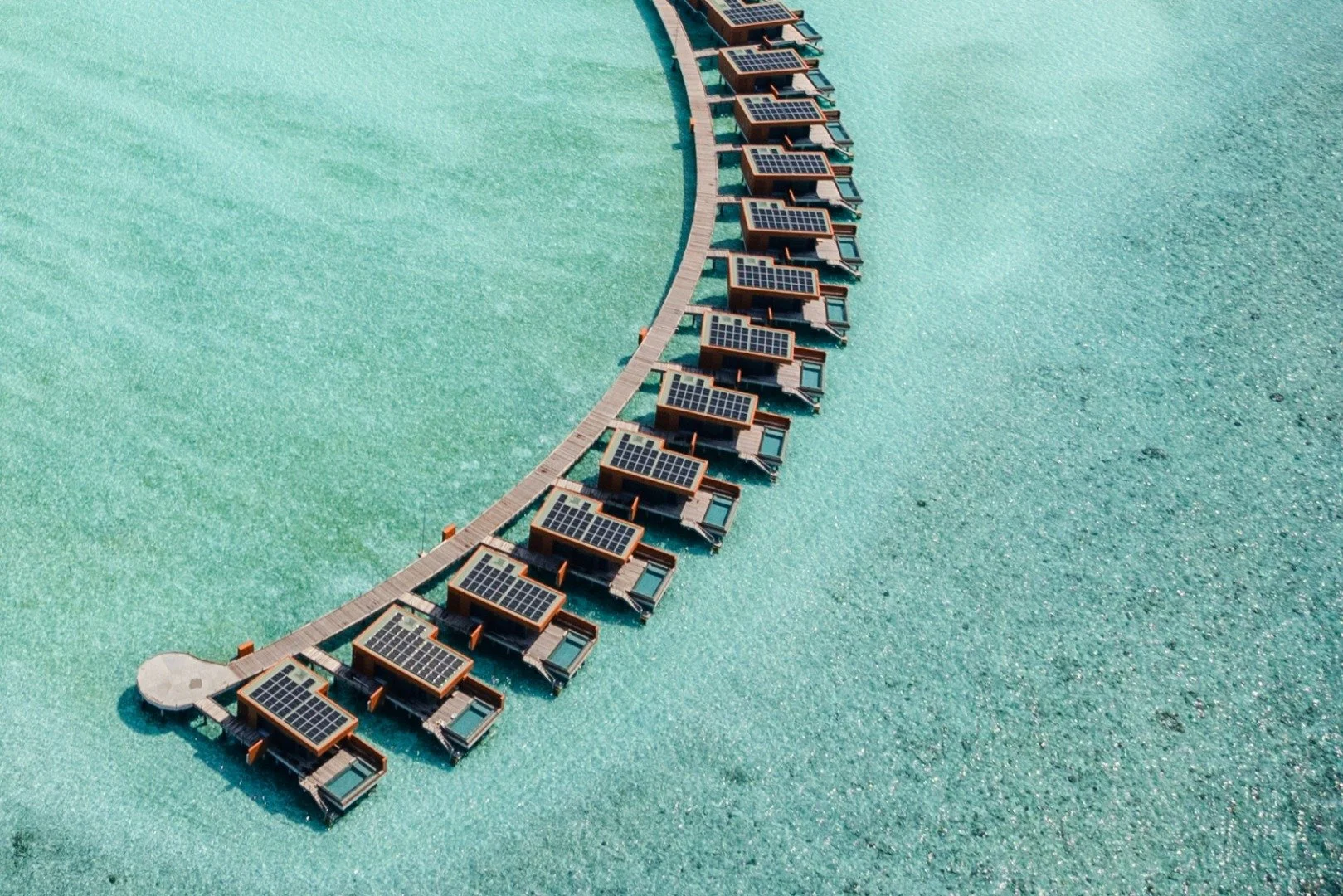 Luftbild av lyxiga vattenvillor med solpaneler över turkost vatten vid en brygga.