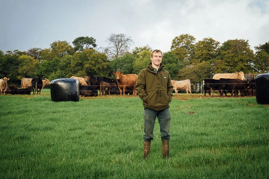 Chris stood in grass field with cattle and bales of haylage behind him