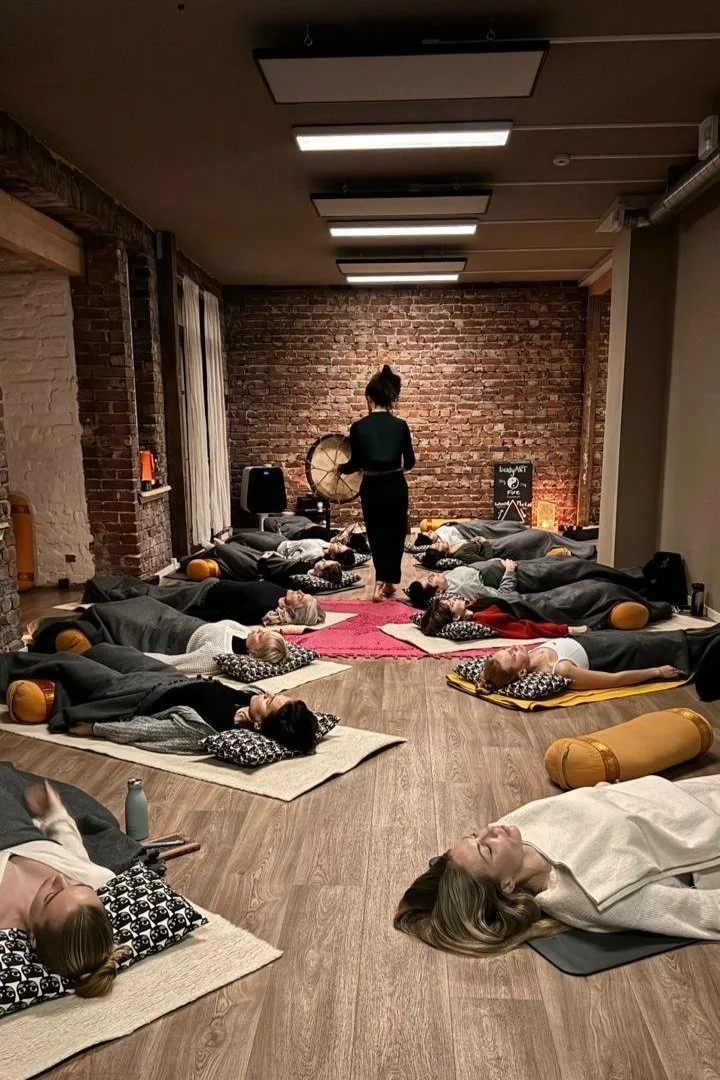 Shows a group of people who are very comfortable relaxed and laying down with Judi holding shamanic drum guiding them through a meditation after a cacao ceremony