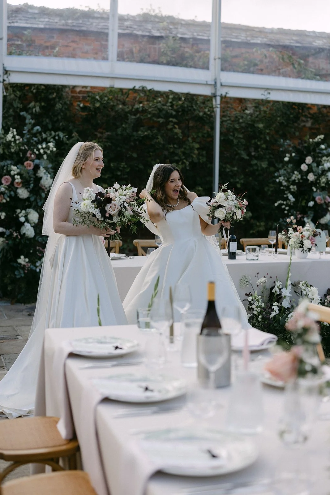 brides looking in excitement at the reveal of their wedding breakfast at Kingston Estate. Natural and elegant table flowers with meadow arrangements and broken arch dressing the top table