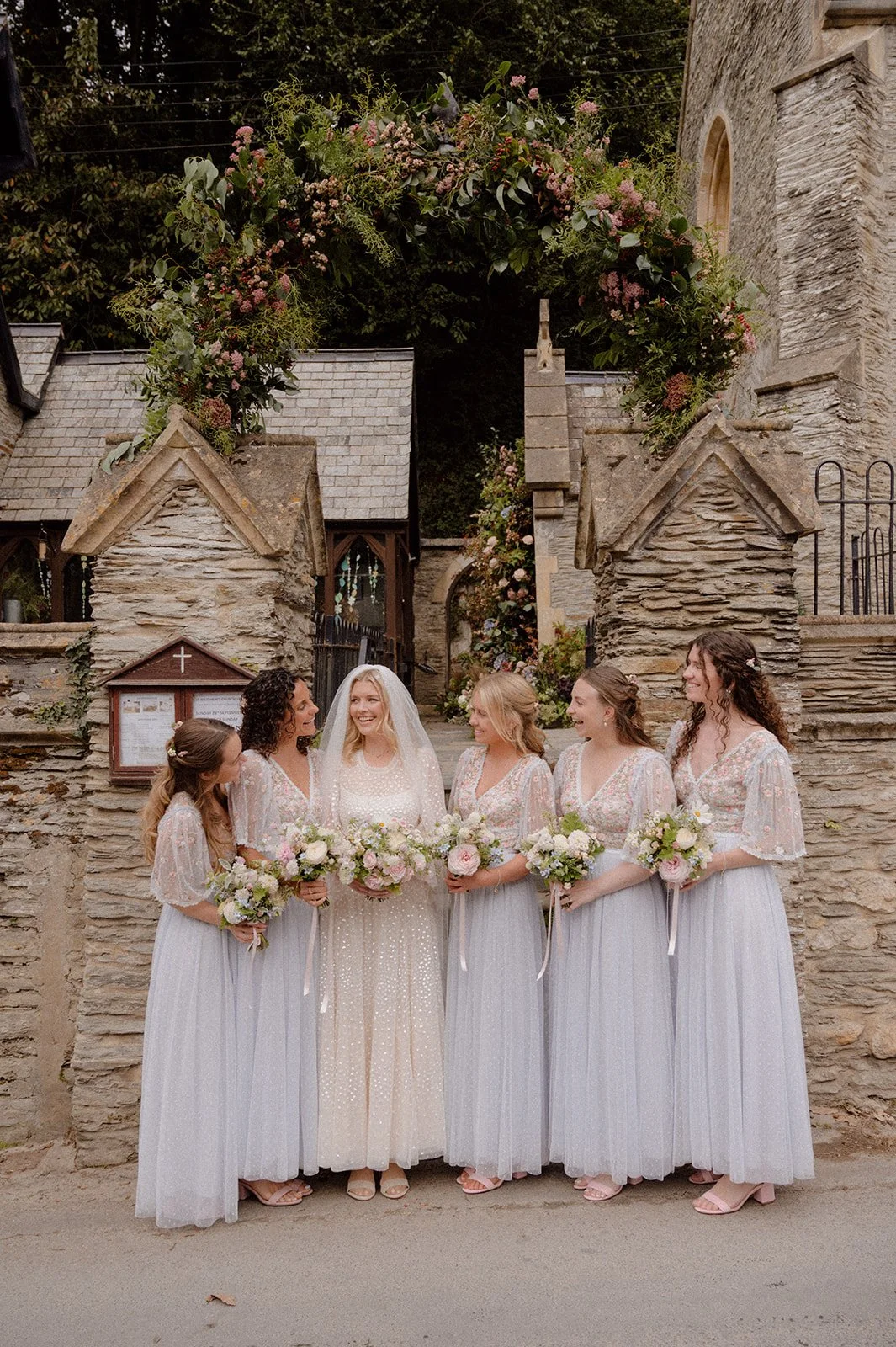 bride and bridesmaids with summer bouquets by flower arch at Lee Bay Church